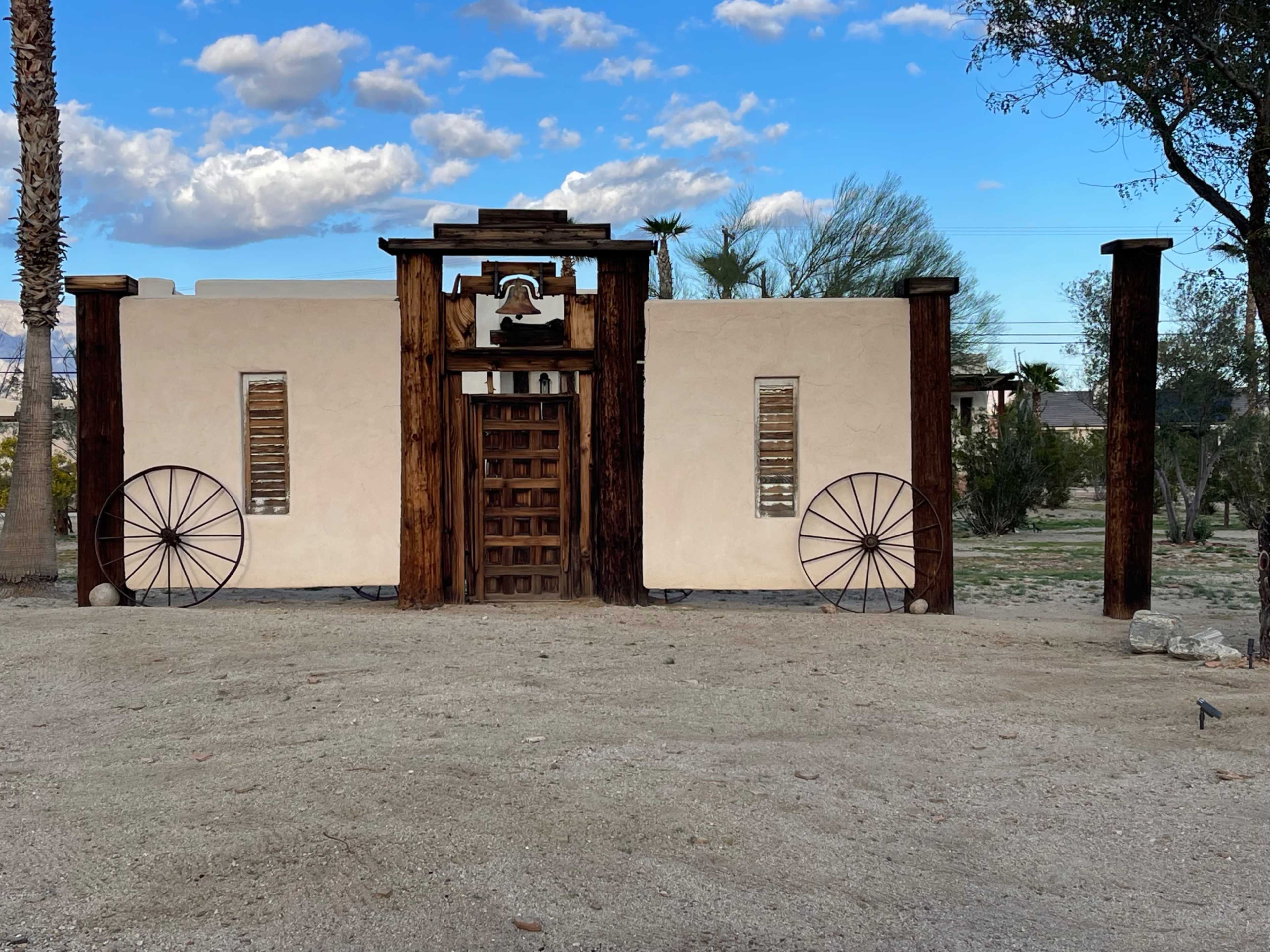 The image shows a rustic adobe-style building with wooden accents, flanked by large wheels and surrounded by sparse desert landscaping.