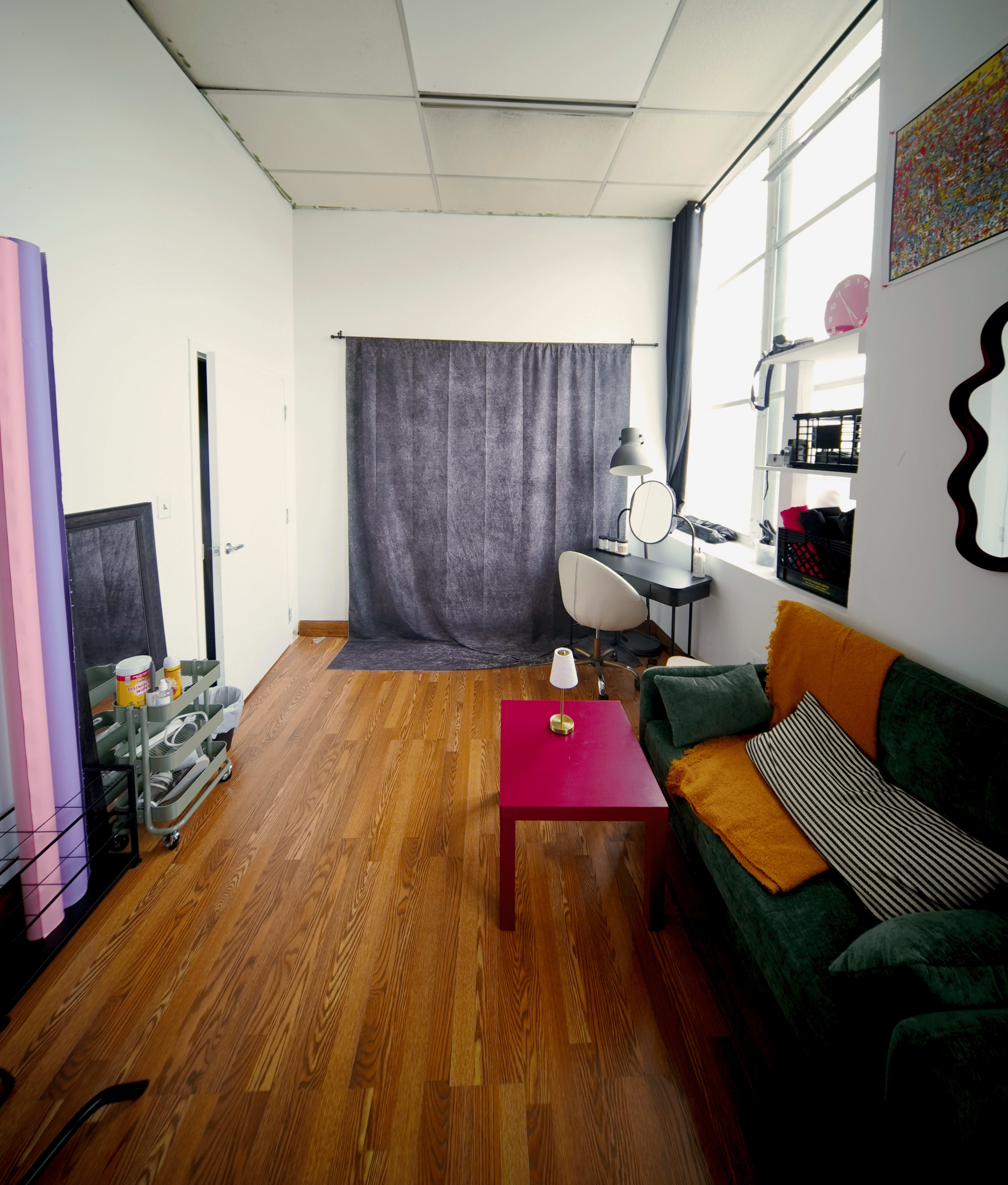The image shows a small studio space featuring a green sofa, a pink coffee table, a photography backdrop, and a chair with a mirror, all positioned against a wall of windows.
