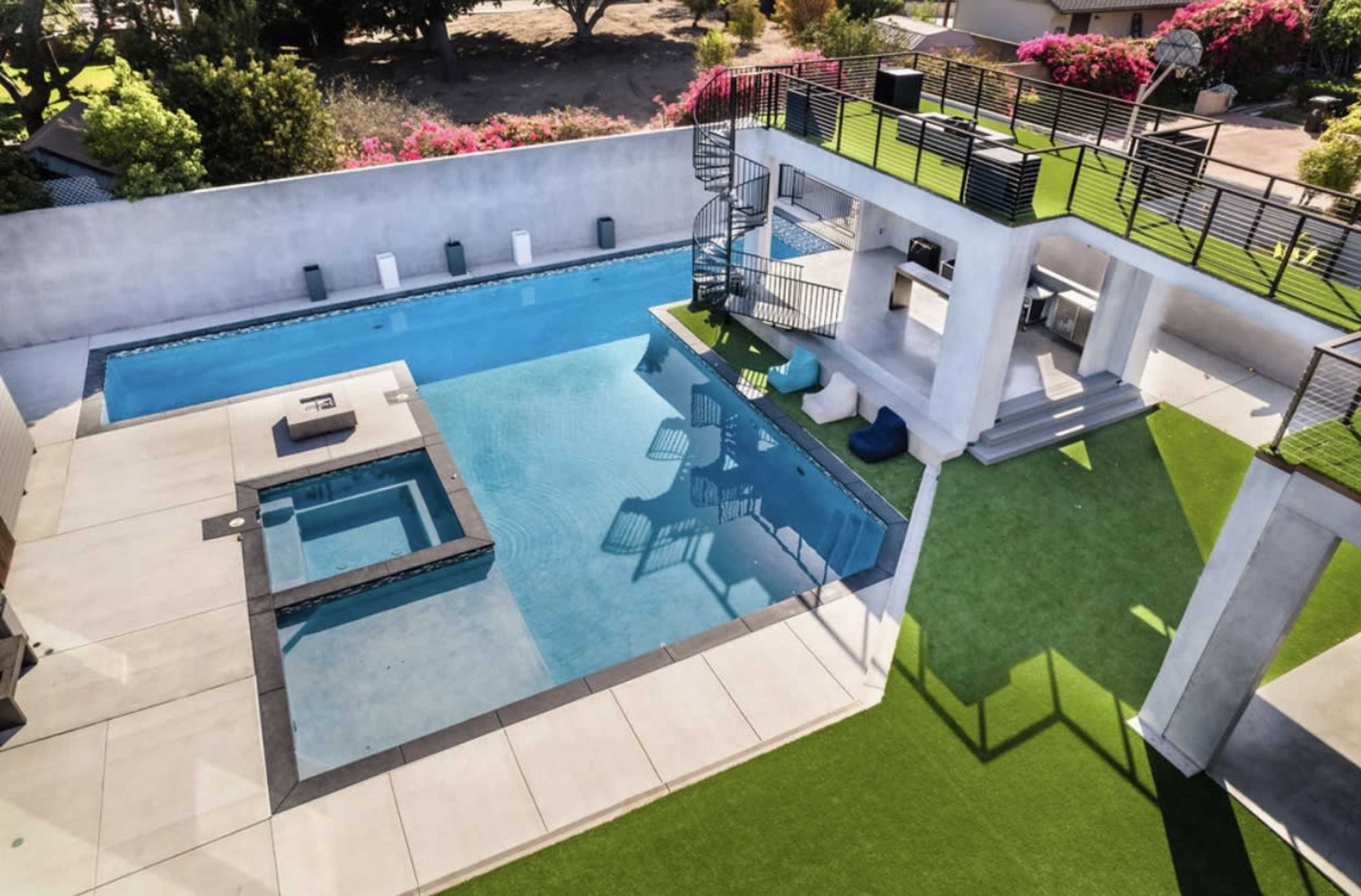 A modern backyard featuring a swimming pool with an attached hot tub, surrounded by a landscaped area with green grass and seating.