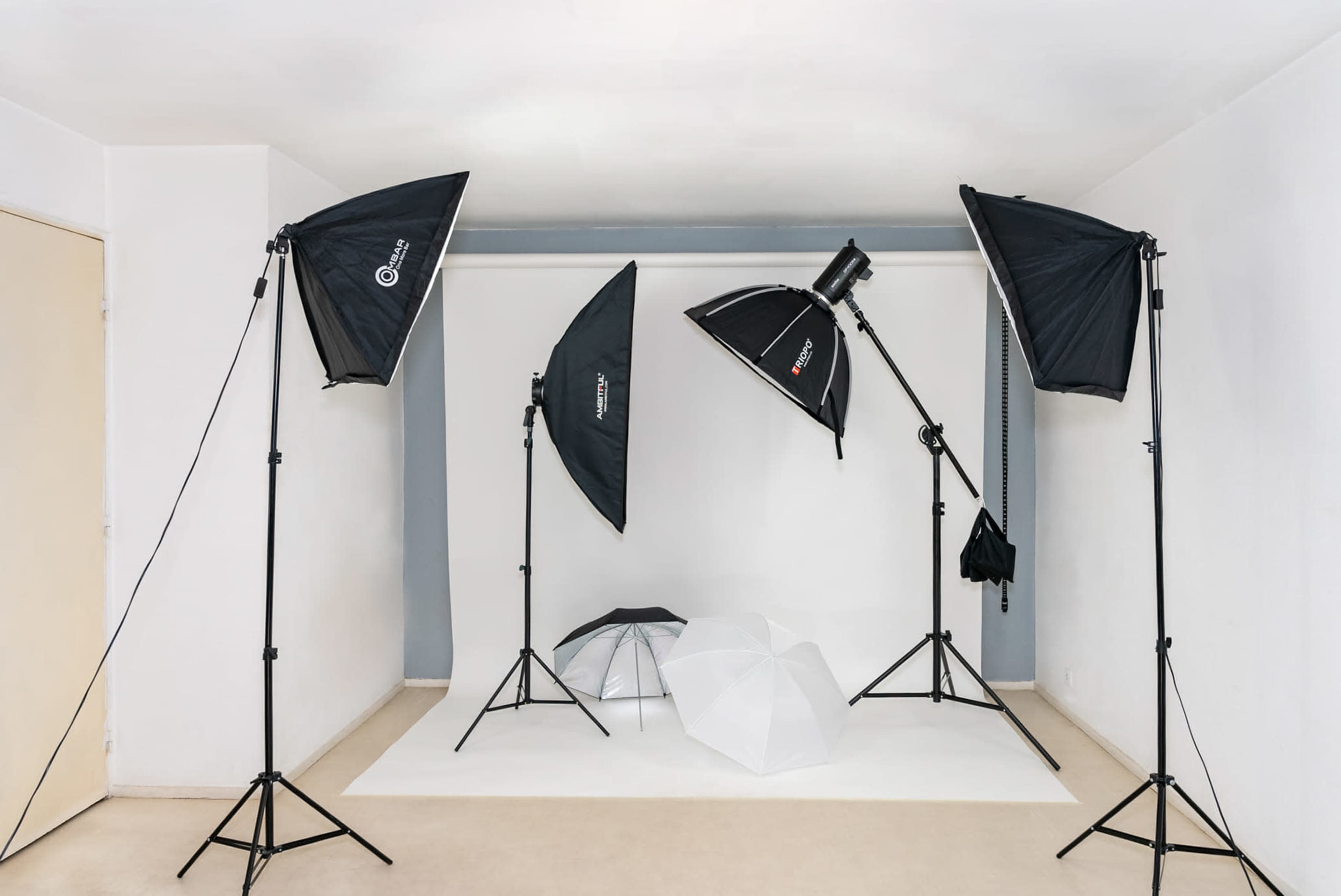 The image shows a photography studio setup with multiple lighting equipment aimed at a plain backdrop and several geometric props on the floor.
