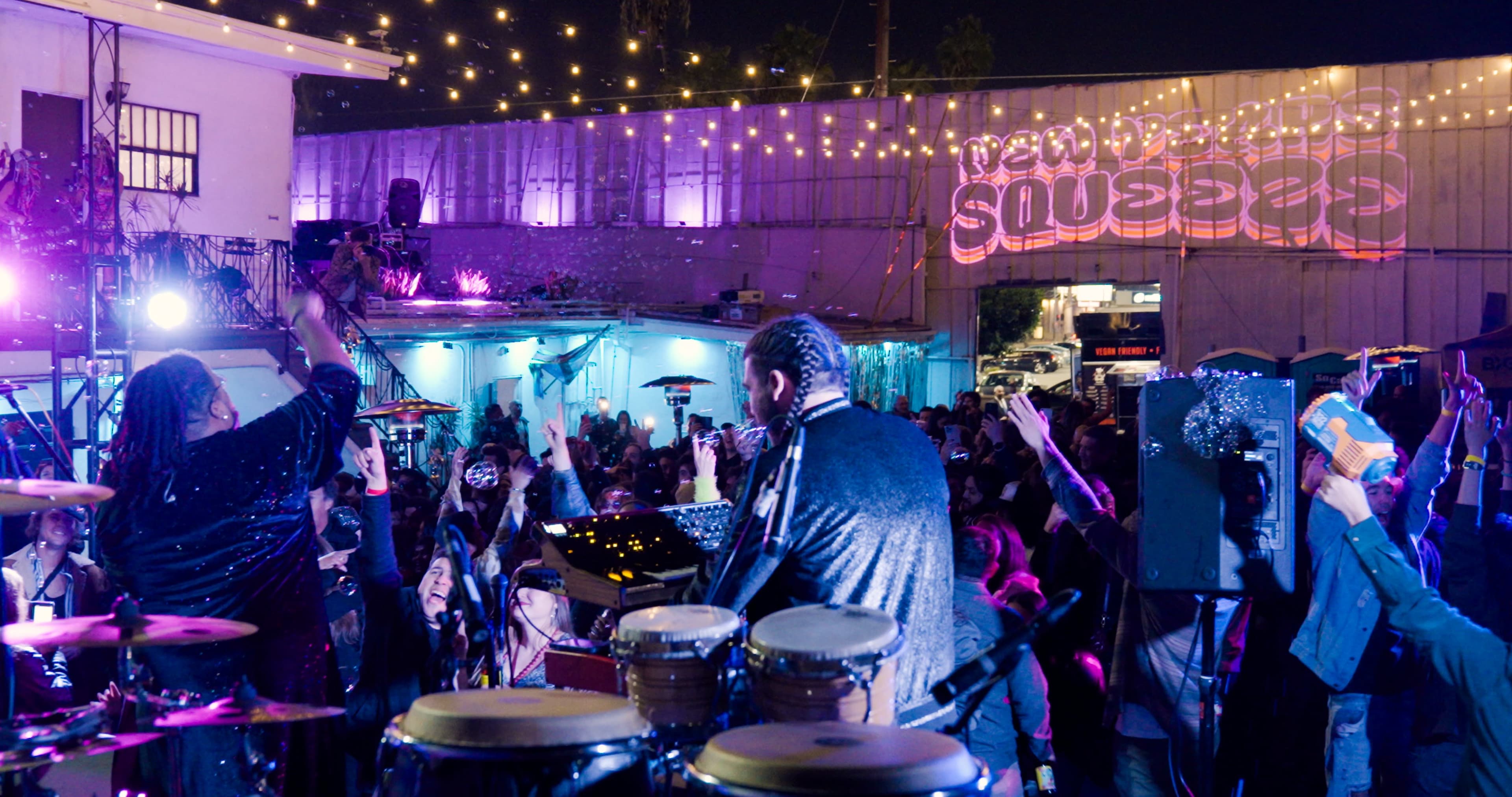 A lively outdoor party features a band performing on stage, surrounded by an energetic crowd and decorative string lights.