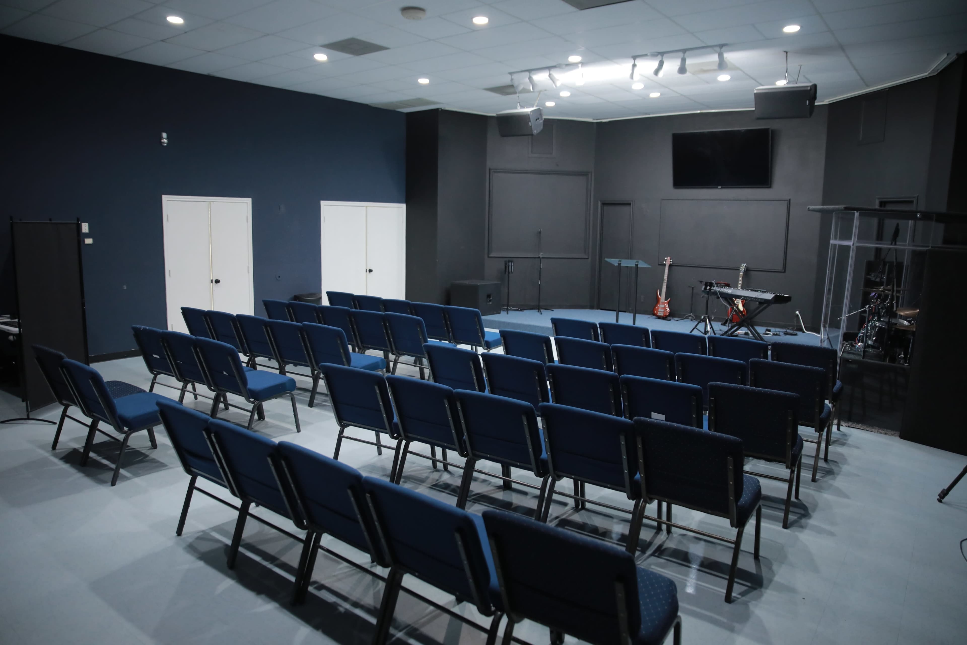 A sparsely arranged indoor space with rows of blue chairs facing a stage that has musical instruments set up.