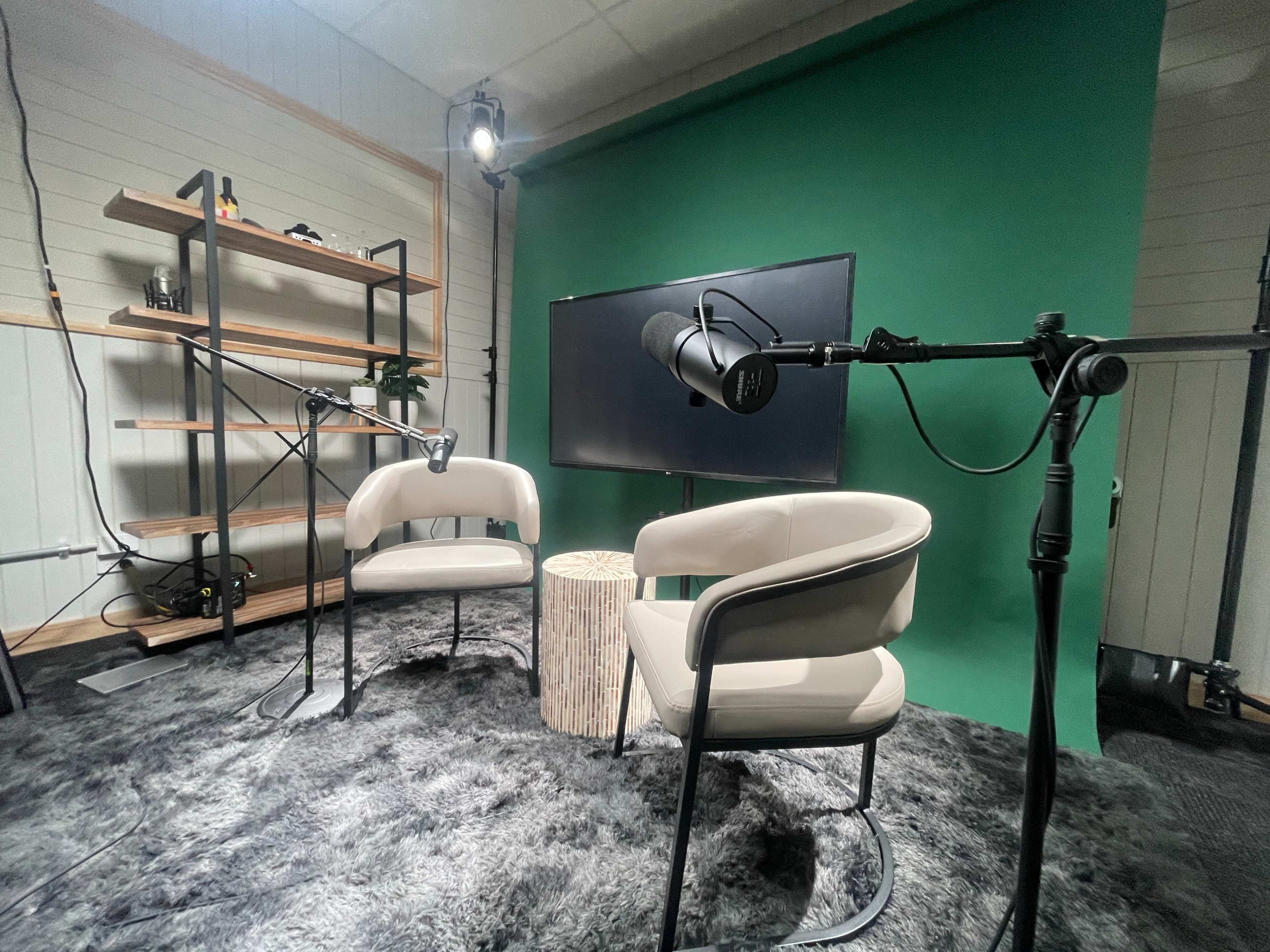 The image shows a podcast recording setup with two chairs, a small table, microphones on stands, a television, and a green backdrop.