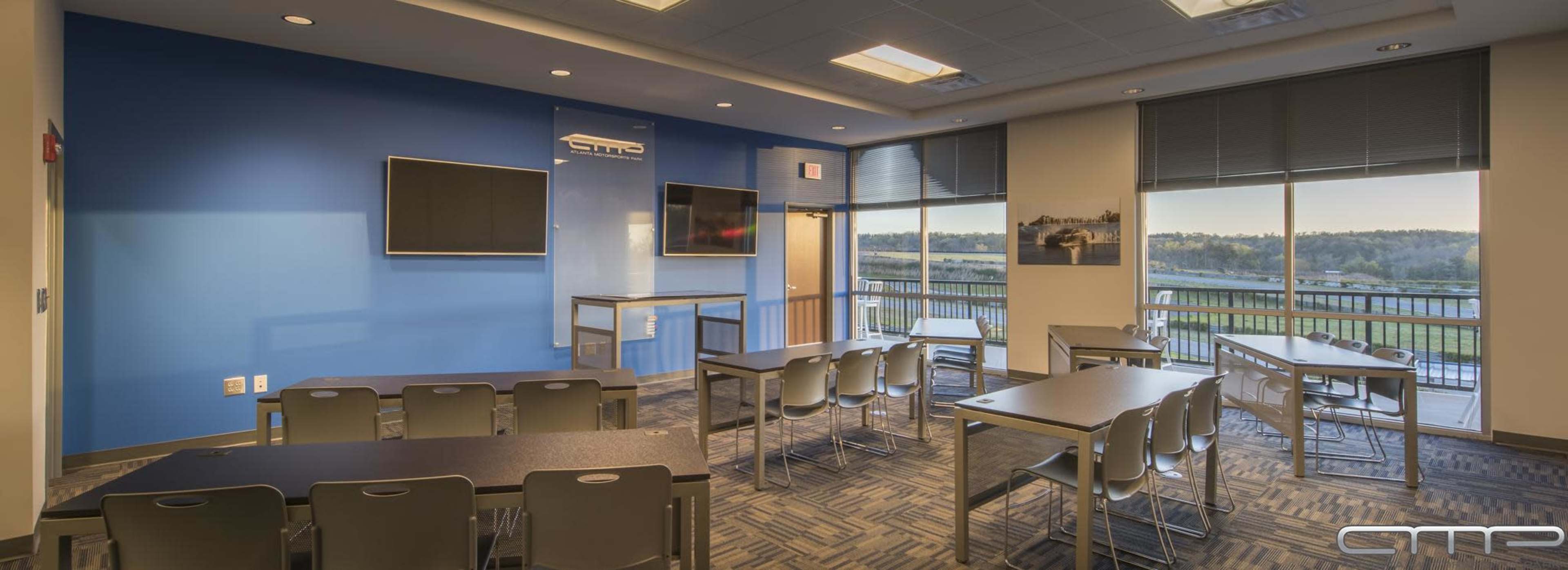 The image shows a modern classroom with rows of tables and chairs, large windows offering a view, and two wall-mounted screens.
