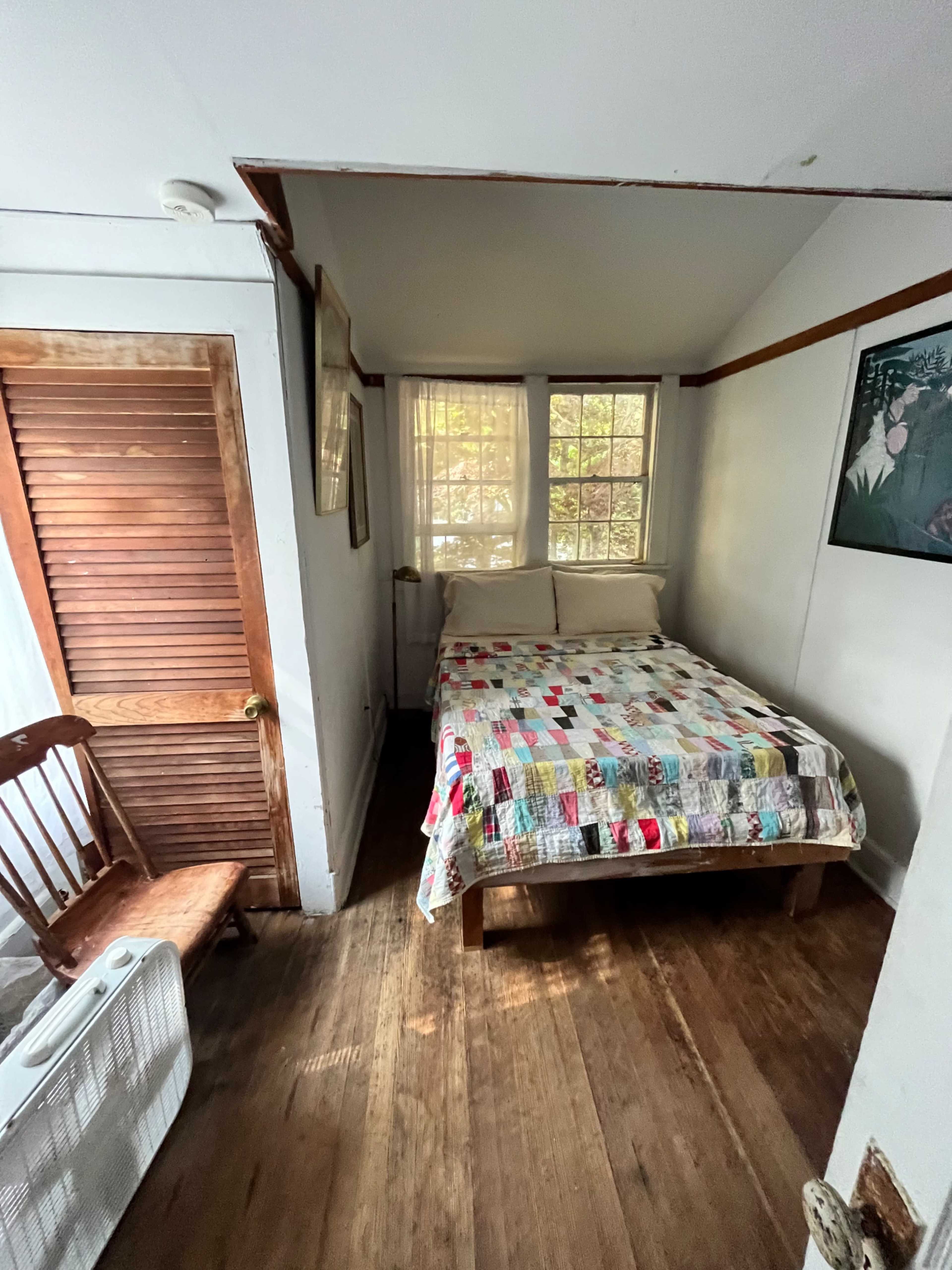 The image shows a small bedroom featuring a bed with a colorful quilt, a wooden chair, and a window allowing natural light to enter.