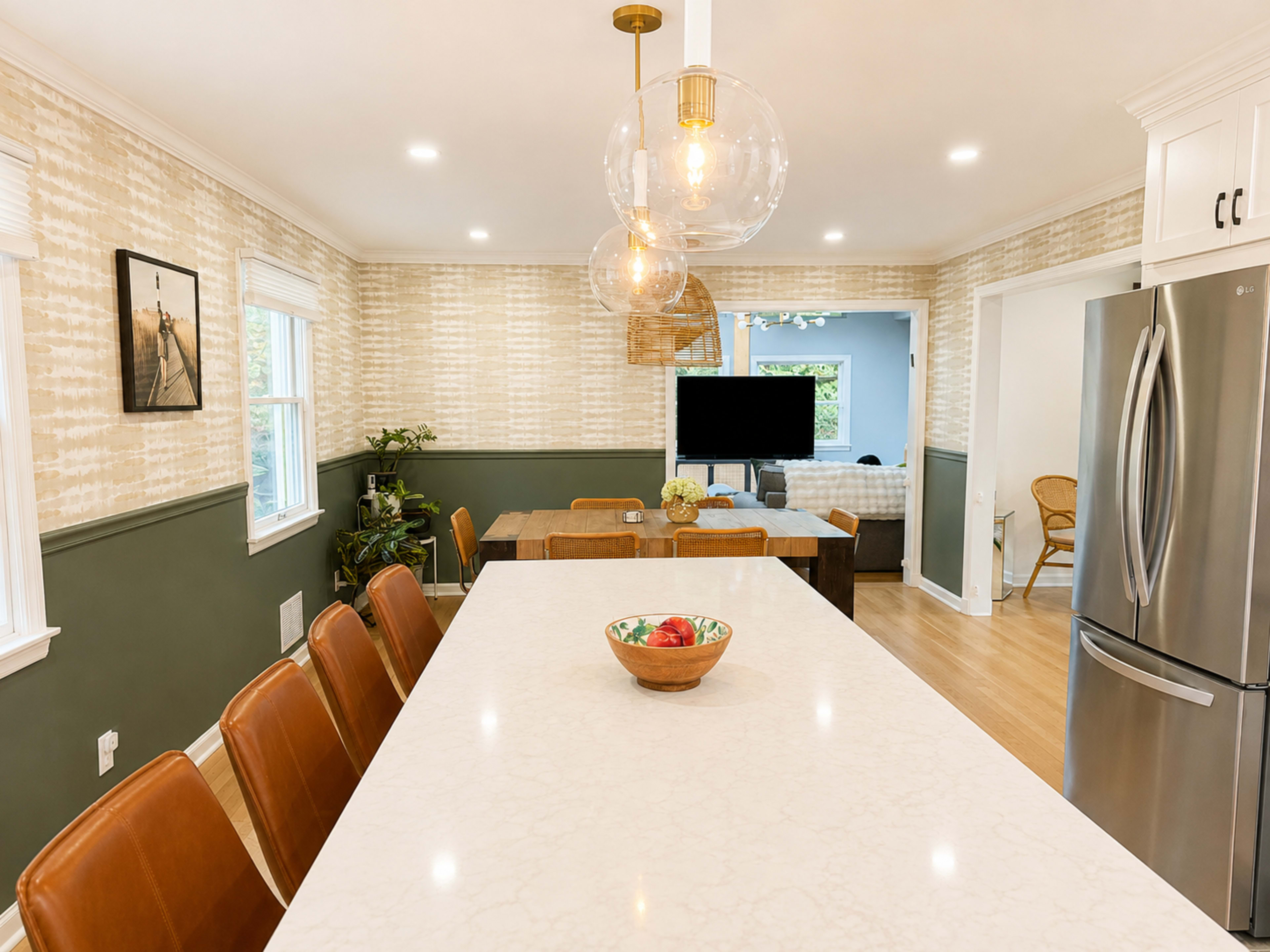 A modern kitchen features a large island with seating, pendant lights, and a dining table, bordered by patterned wallpaper and a living area in the background.