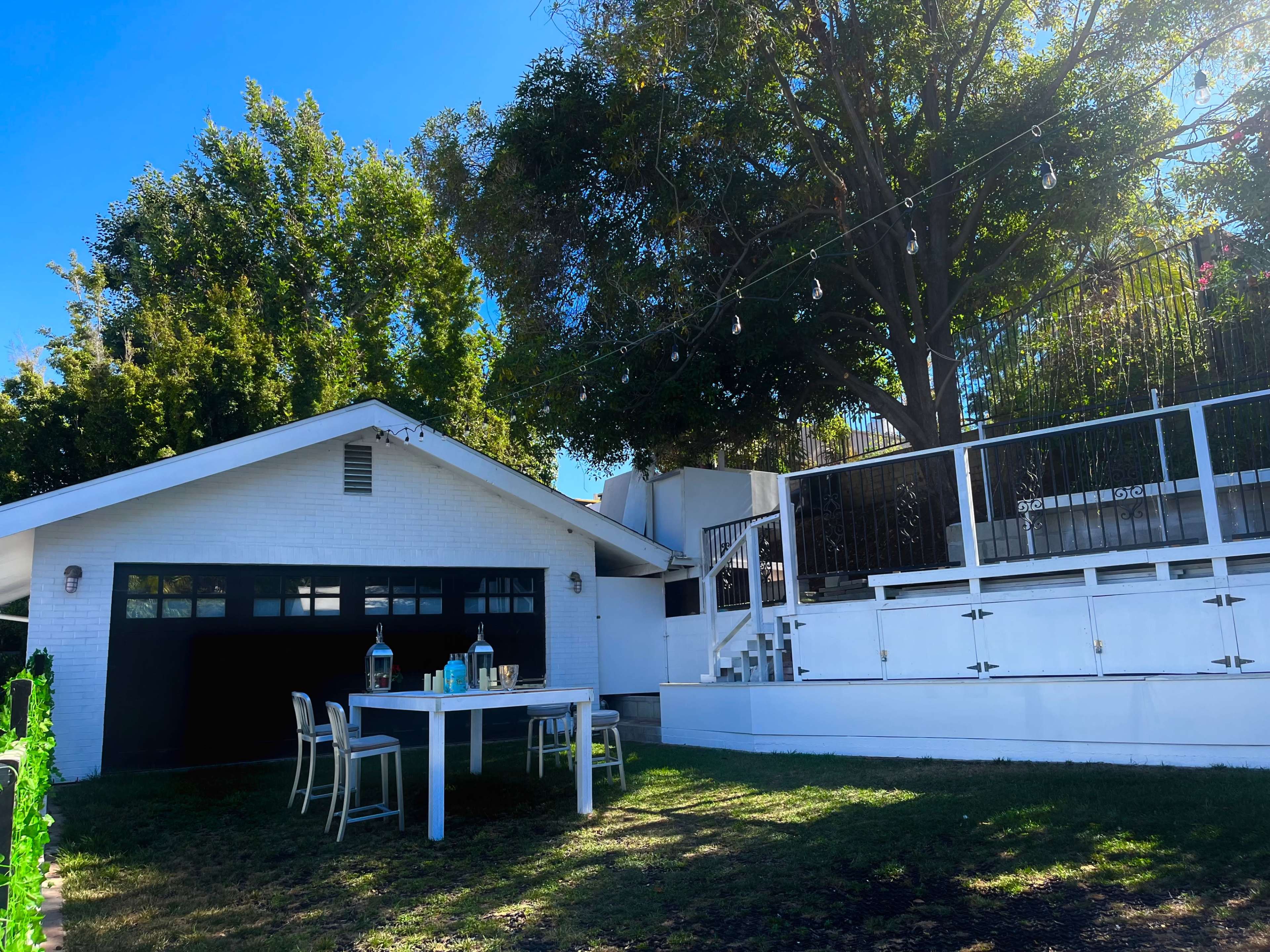 A white house with a garage sits beside a patio area featuring a table and chairs beneath a large tree.