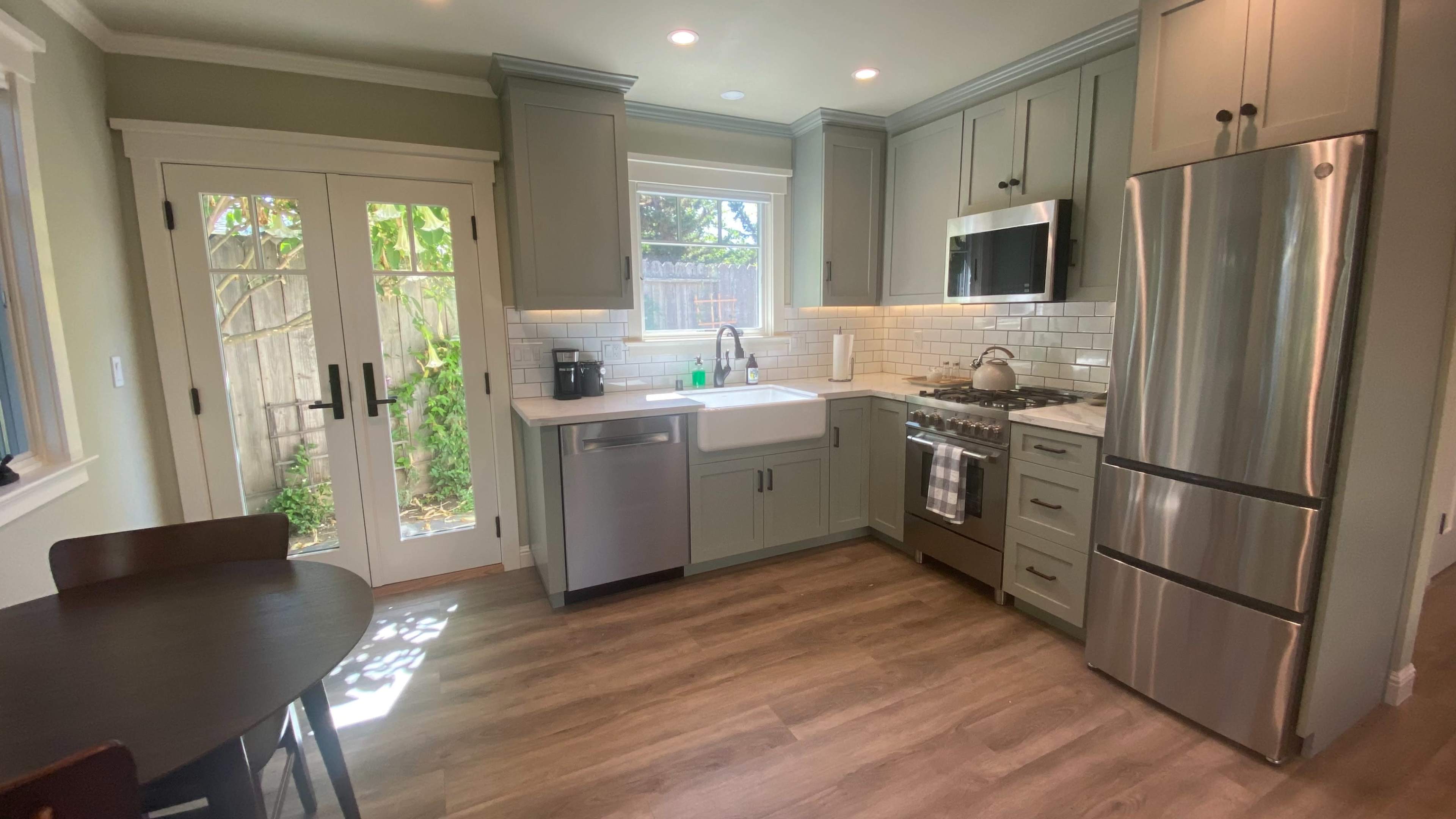 The image shows a modern kitchen with gray cabinets, stainless steel appliances, a white farmhouse sink, and a small dining table.