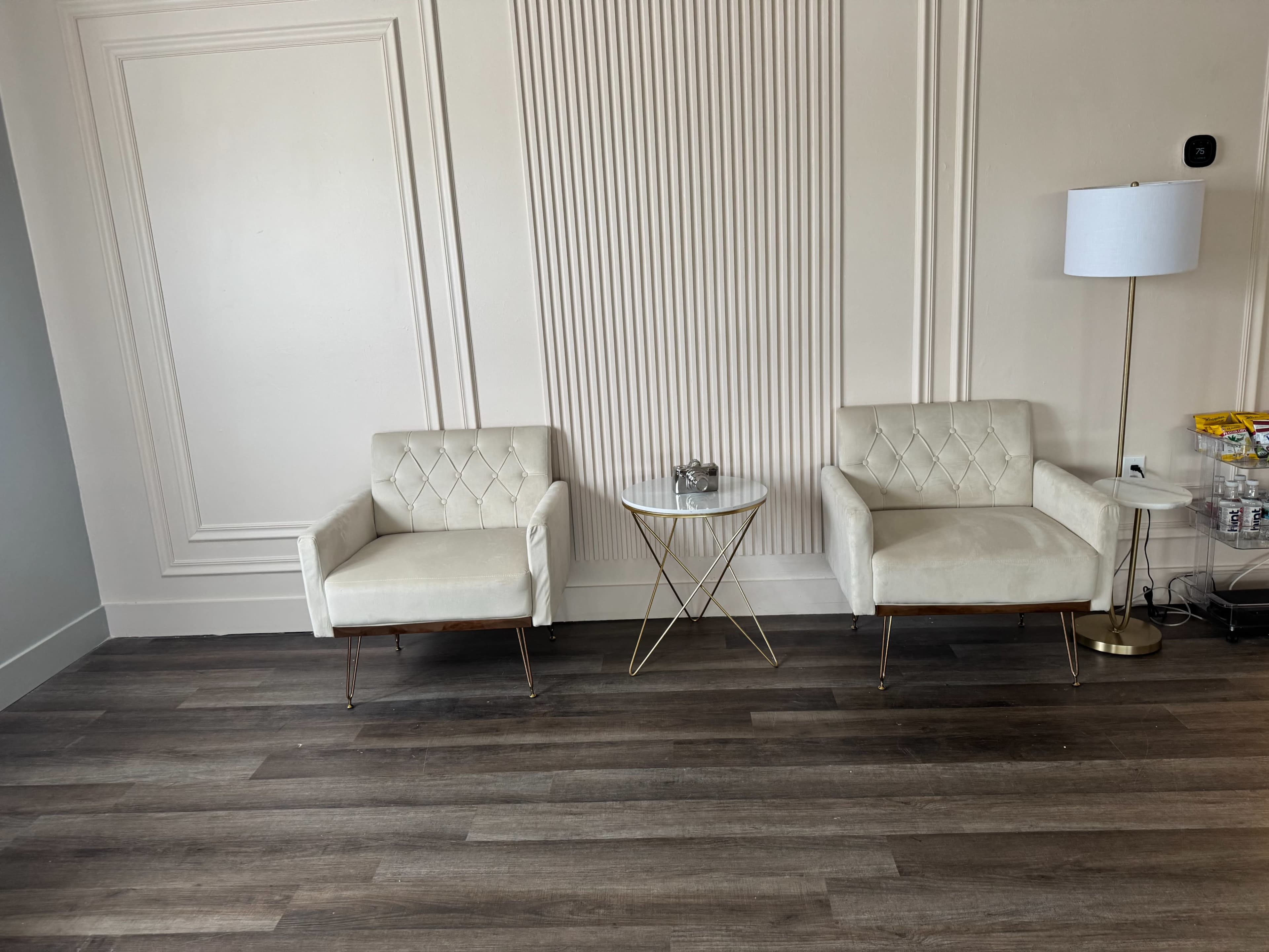 Two white armchairs are positioned opposite each other beside a small golden side table in a room with light-colored walls and wooden flooring.