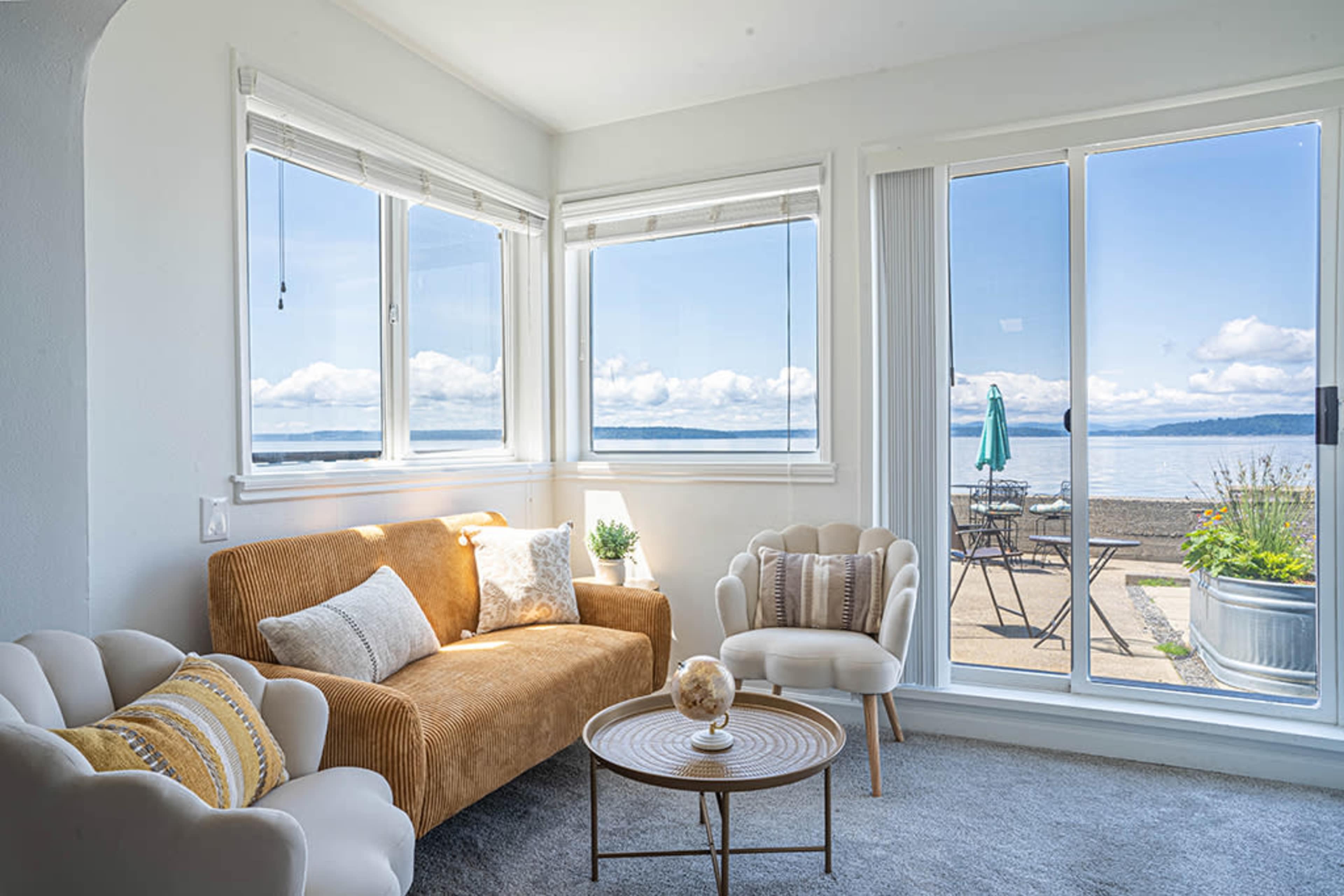 A cozy living room features a brown couch and two white chairs with large windows overlooking a waterfront patio.