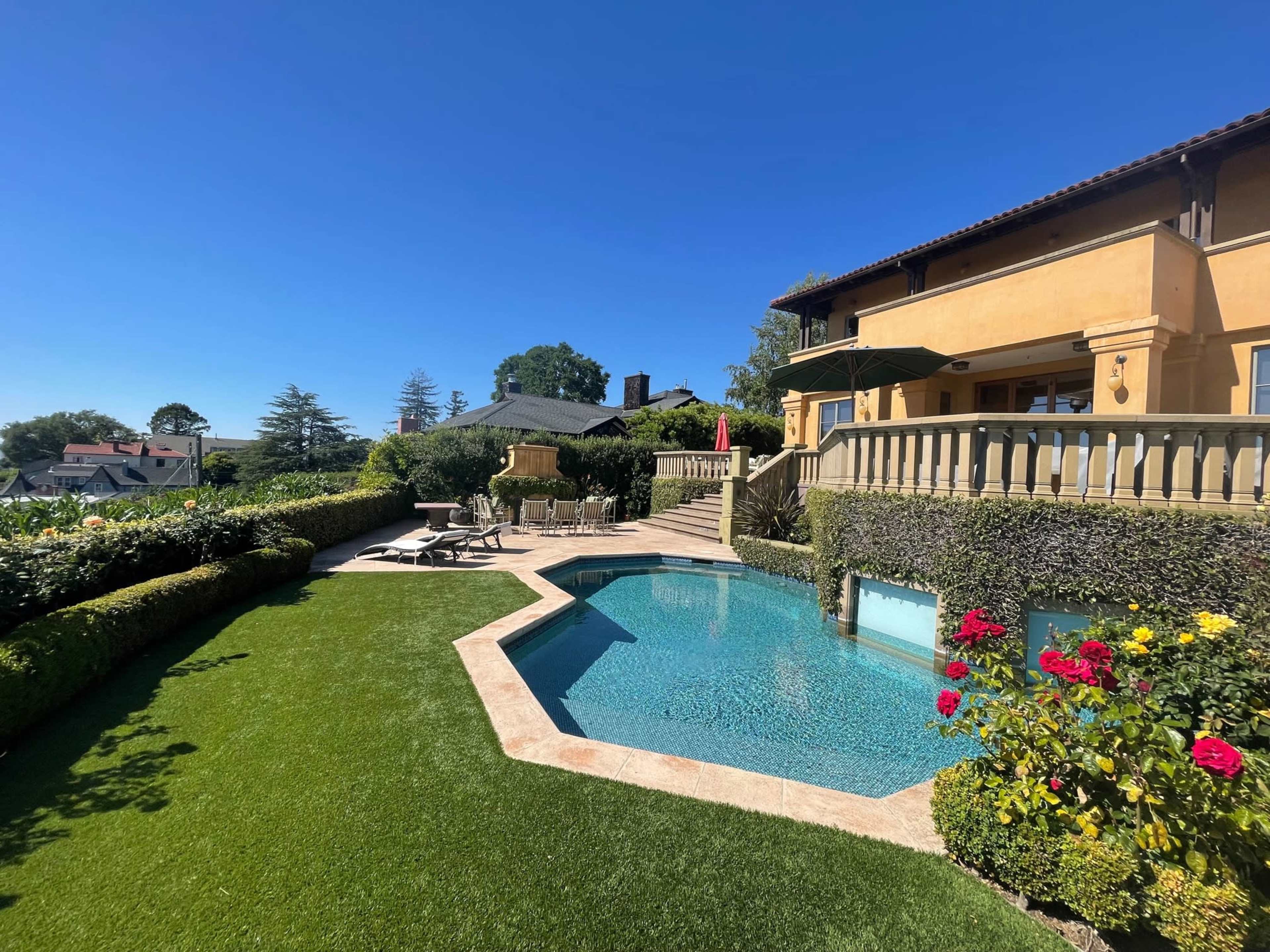 The image shows a residential property with a landscaped yard featuring a swimming pool, lounge chairs, and flower bushes under a clear blue sky.