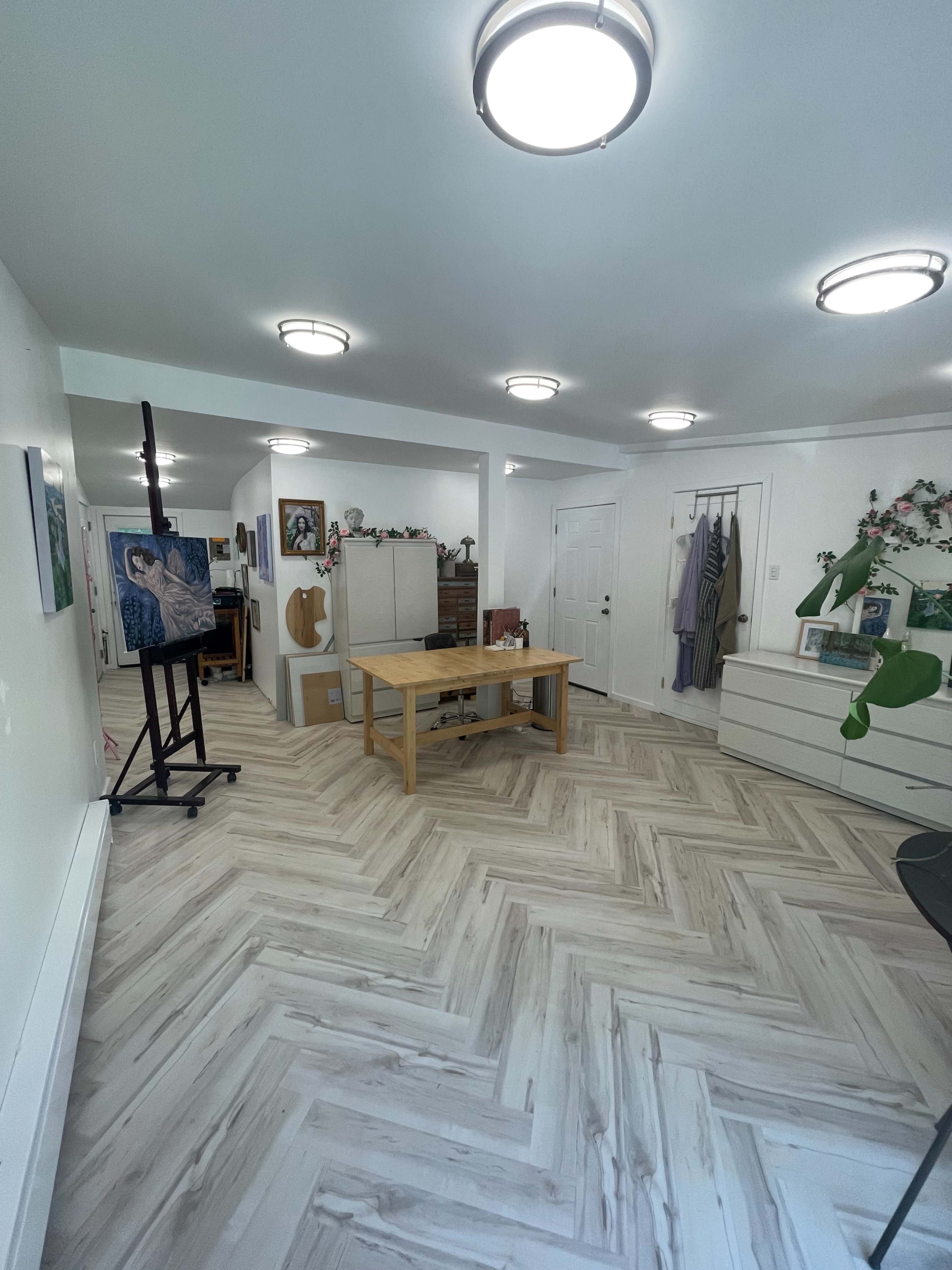 The image shows a brightly lit room with a wooden table, an easel holding a painting, and storage furniture against the walls, all arranged on a chevron-patterned floor.