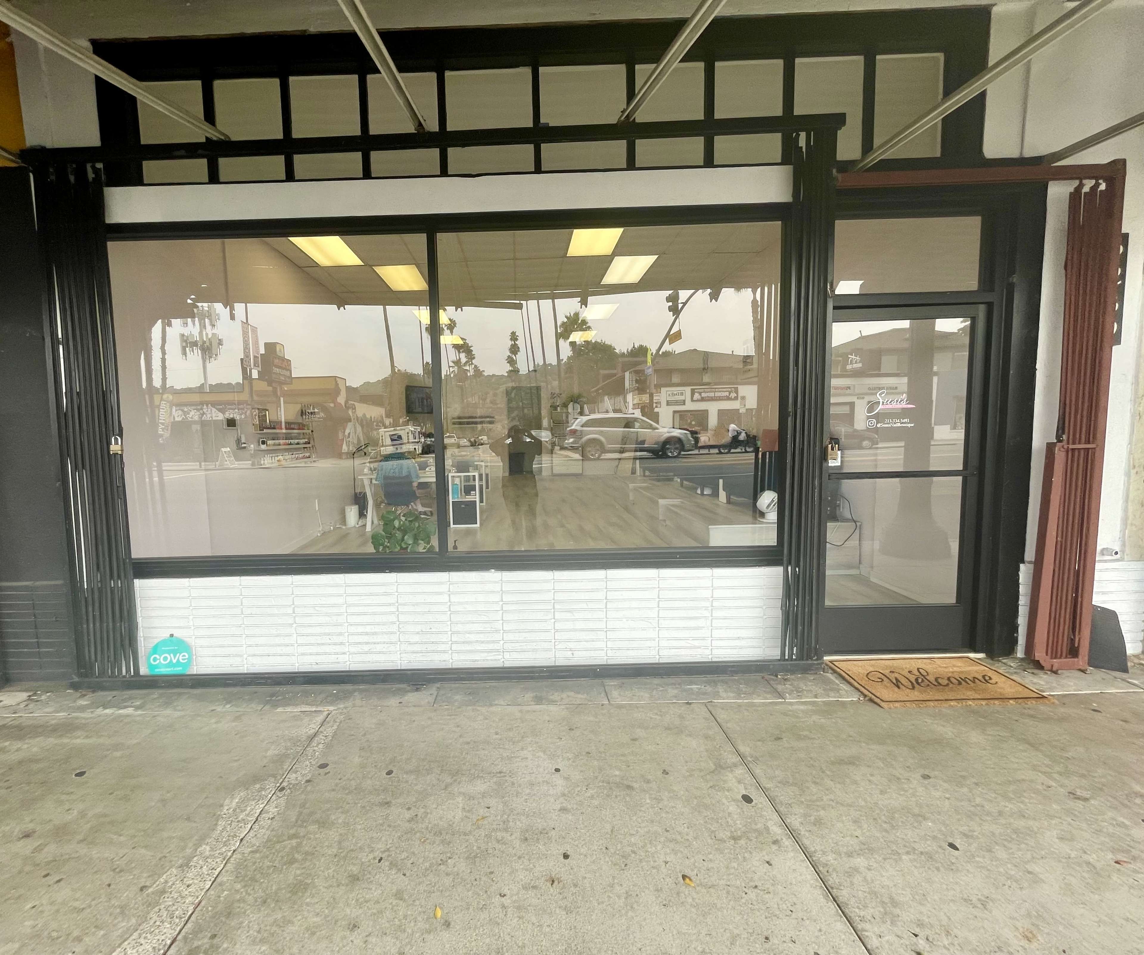 A storefront with large windows displays a welcoming interior featuring seating and decor, along with a "Welcome" mat at the entrance.