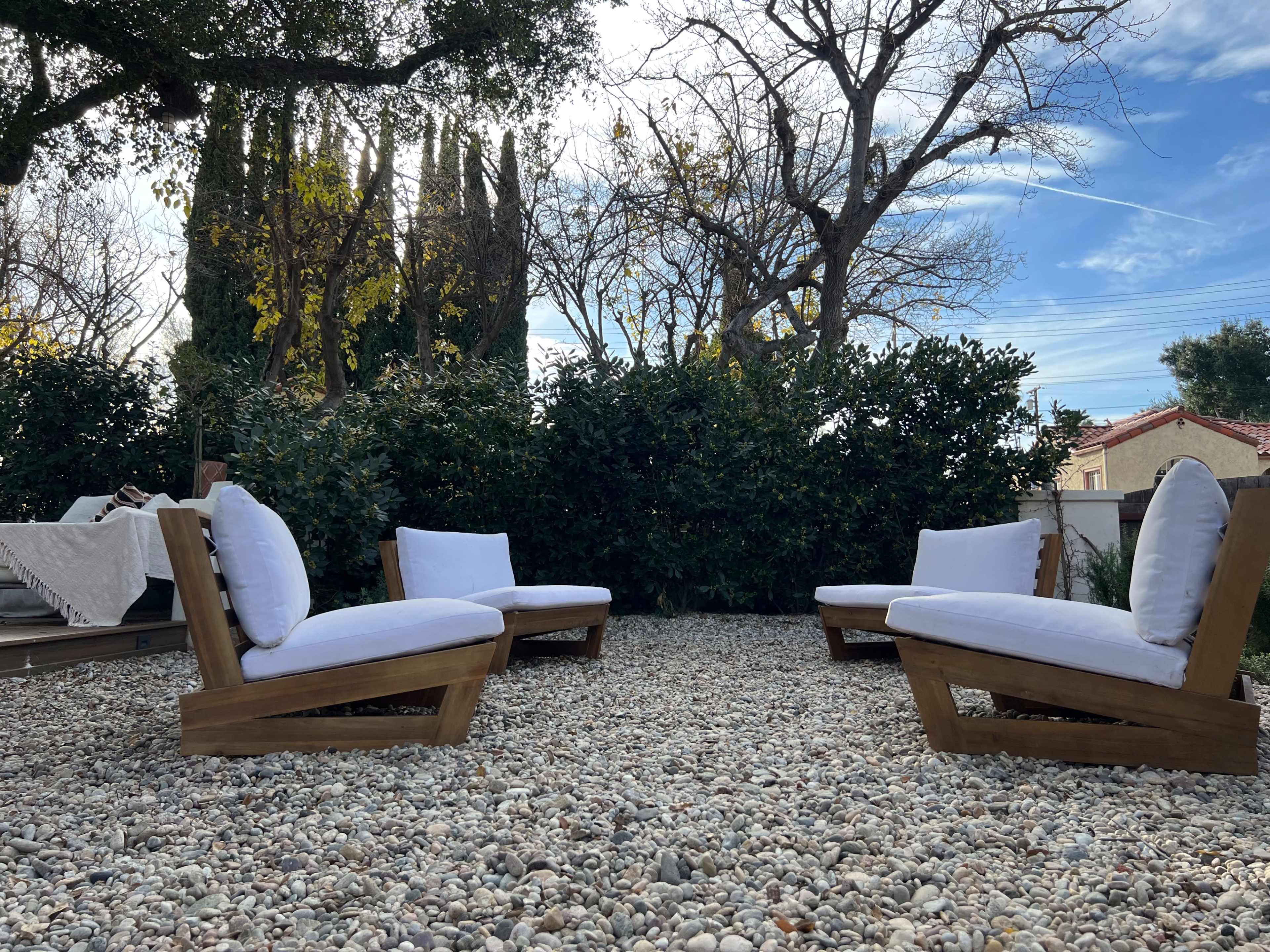 A seating area with four wooden lounge chairs and white cushions is surrounded by gravel and shrubs under a clear sky.