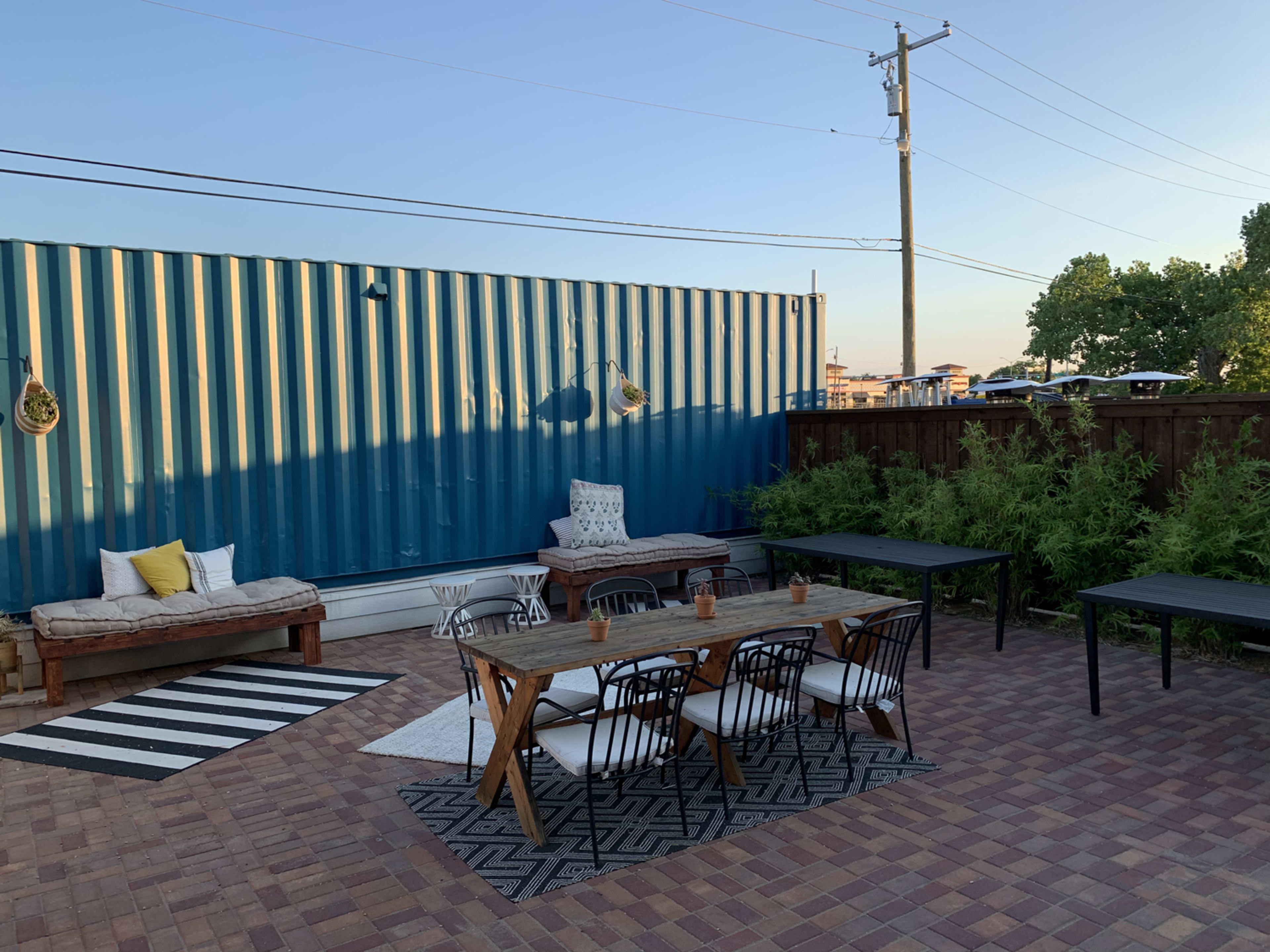 The scene features a patio area with a seating arrangement, a large blue shipping container in the background, and several outdoor tables on a brick surface.