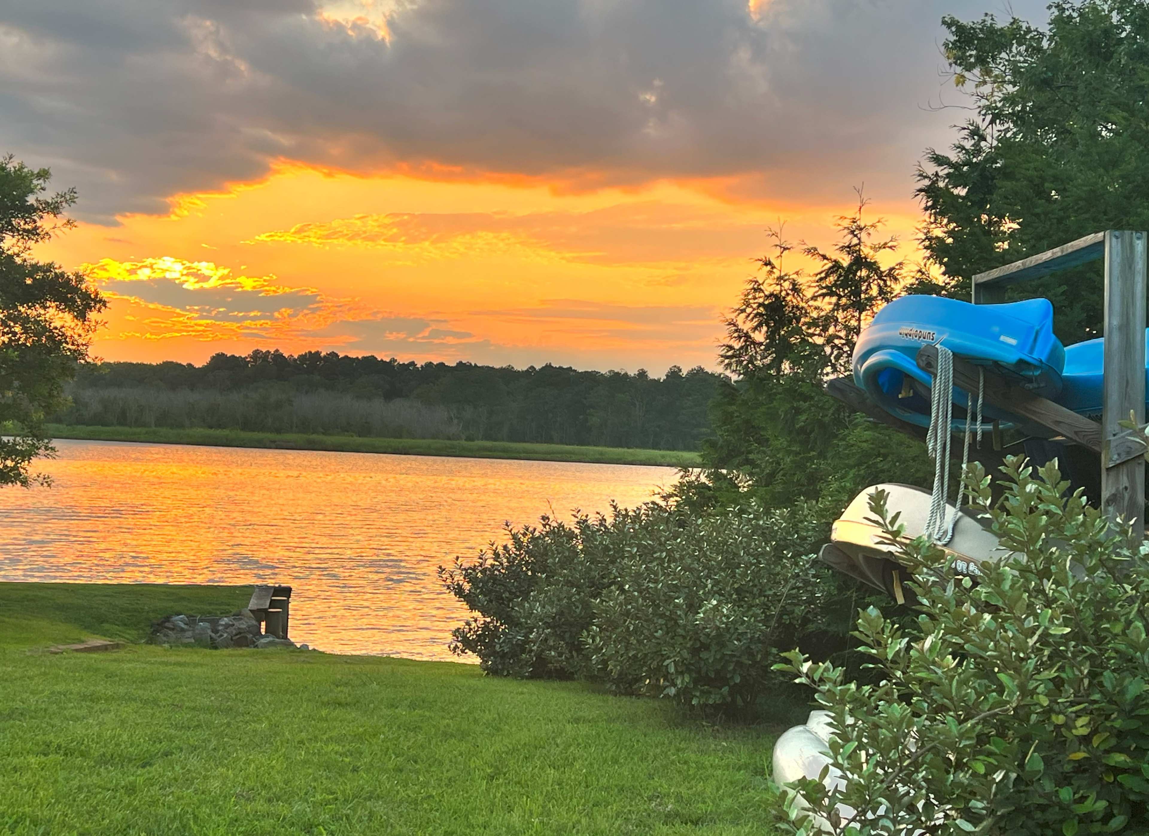 A serene lake at sunset with colorful reflections on the water, surrounded by greenery and kayaks stacked on a wooden structure.