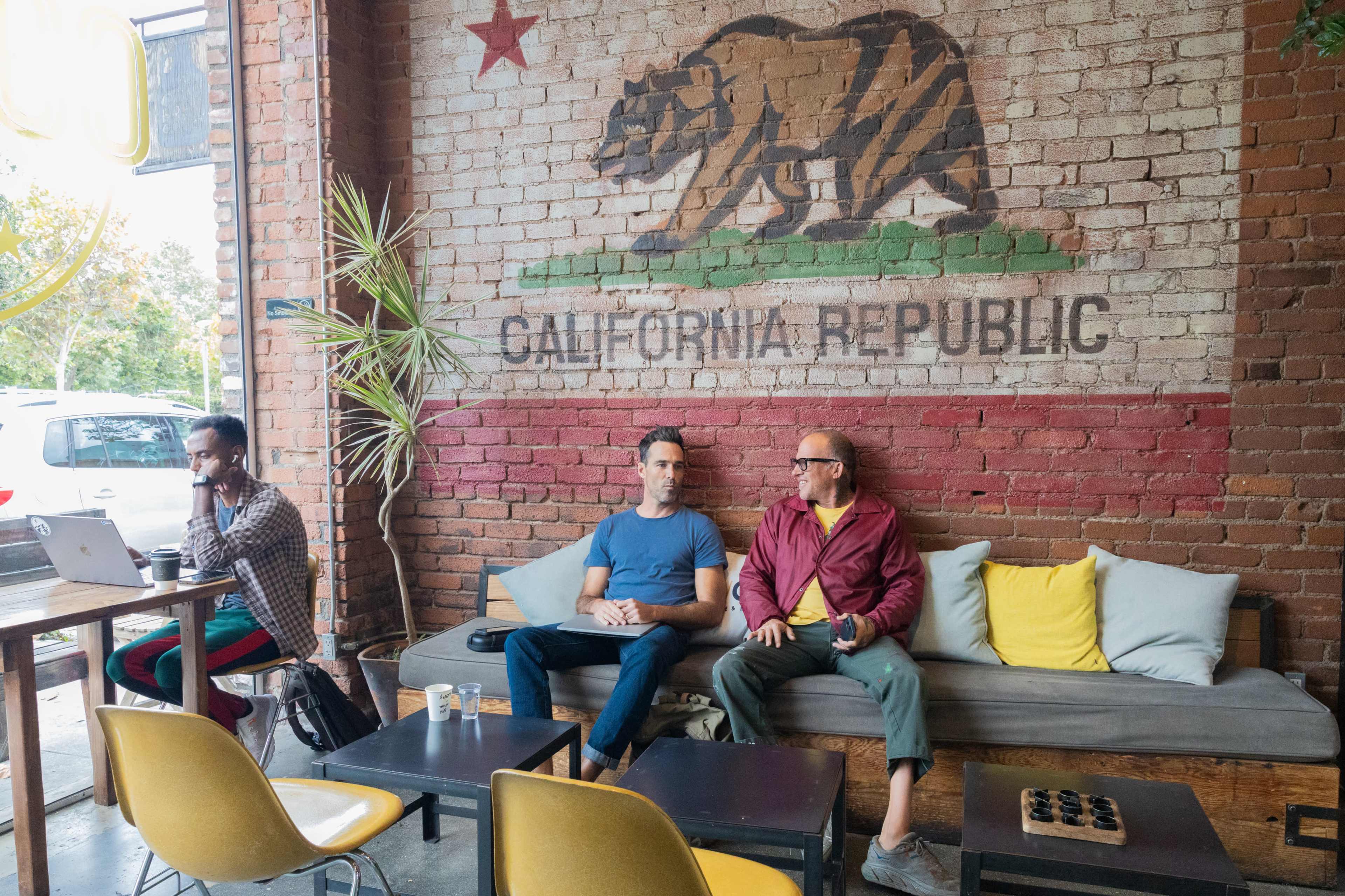 Three men are seated in a cafe with a California Republic mural on the brick wall behind them, while one man works on a laptop at a nearby table.