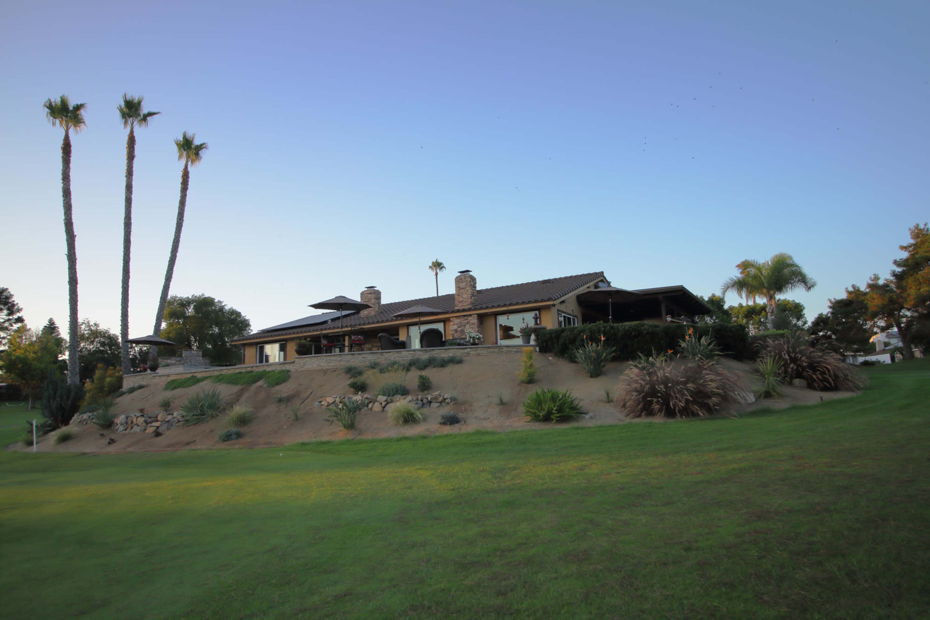 A large house sits on a hillside, surrounded by palm trees and landscaped gardens, with a clear sky above.