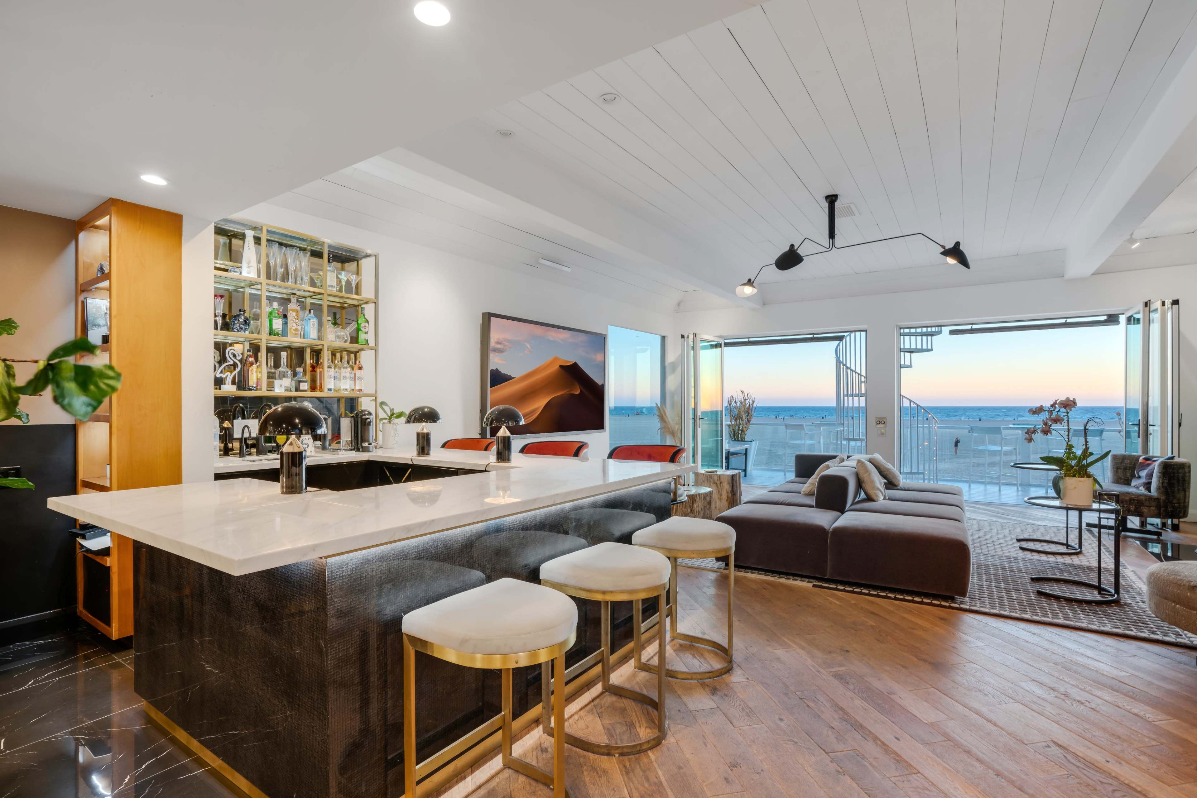 A modern kitchen with a large marble island, featuring a bar area and an ocean view through glass doors.