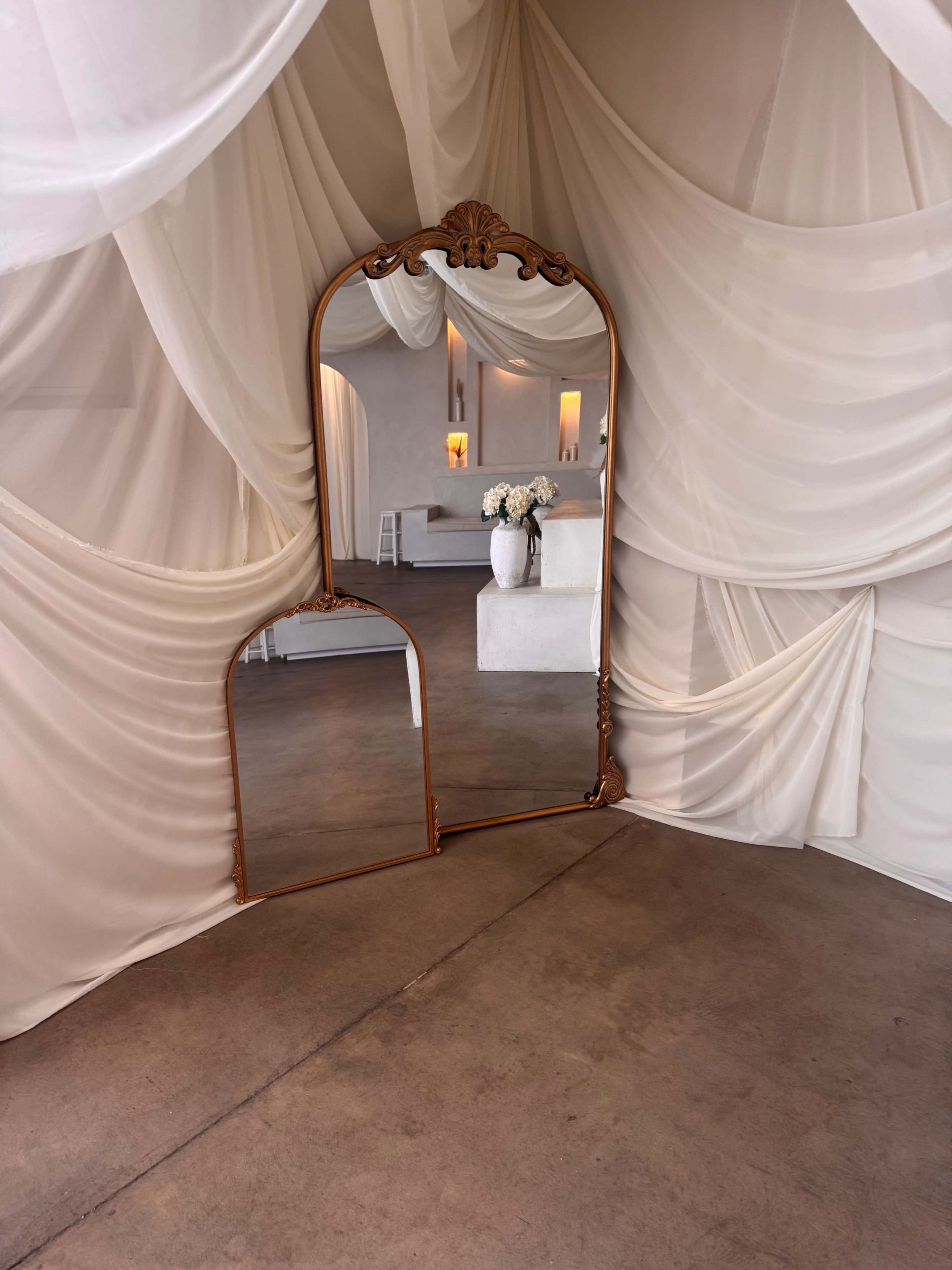The image shows an ornate mirror framed in gold, positioned within a draped fabric setting, reflecting a minimalist interior space with white decor and a floral arrangement.