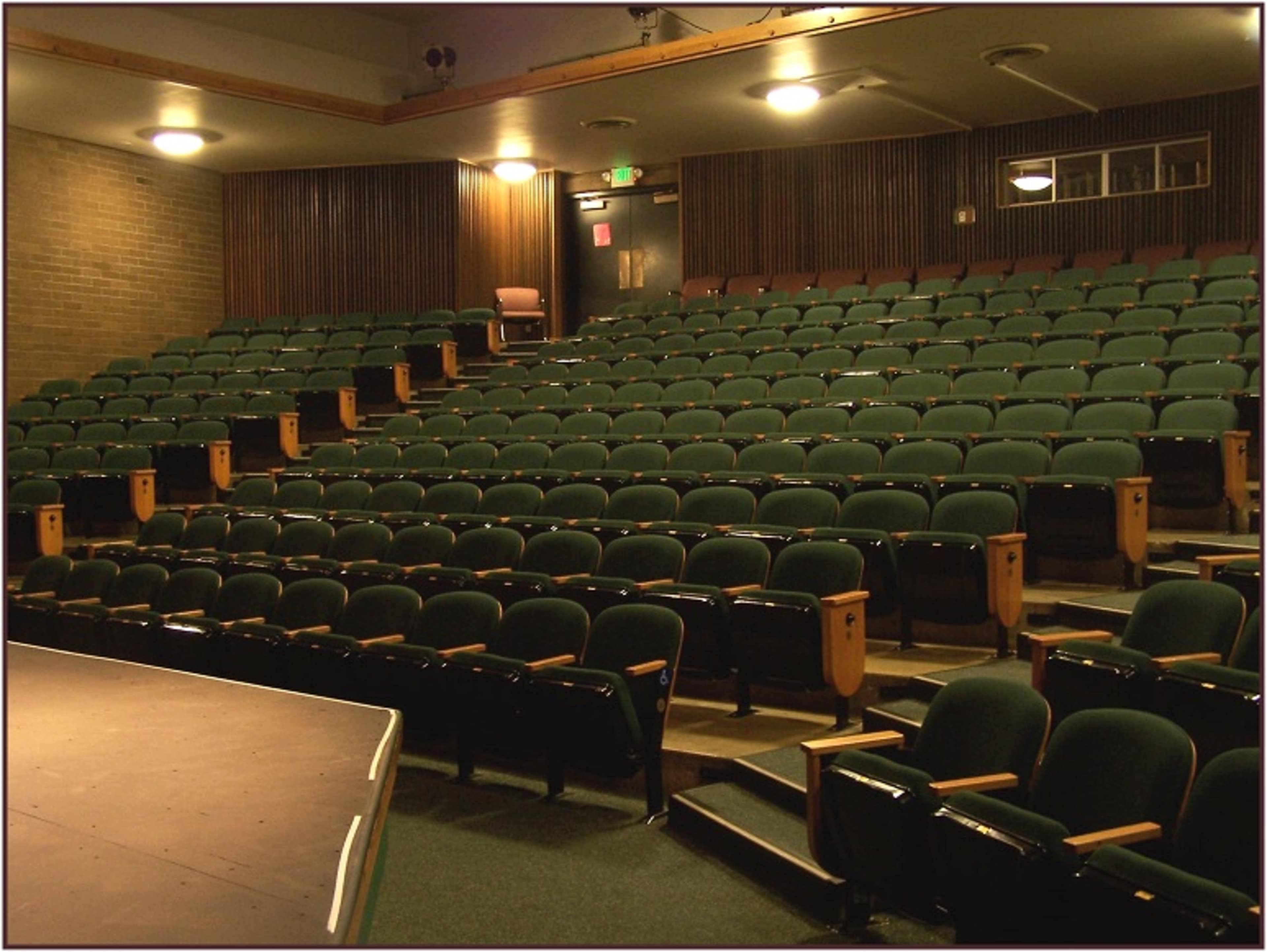 An empty theater with green upholstered seats arranged in rows facing a small elevated stage.