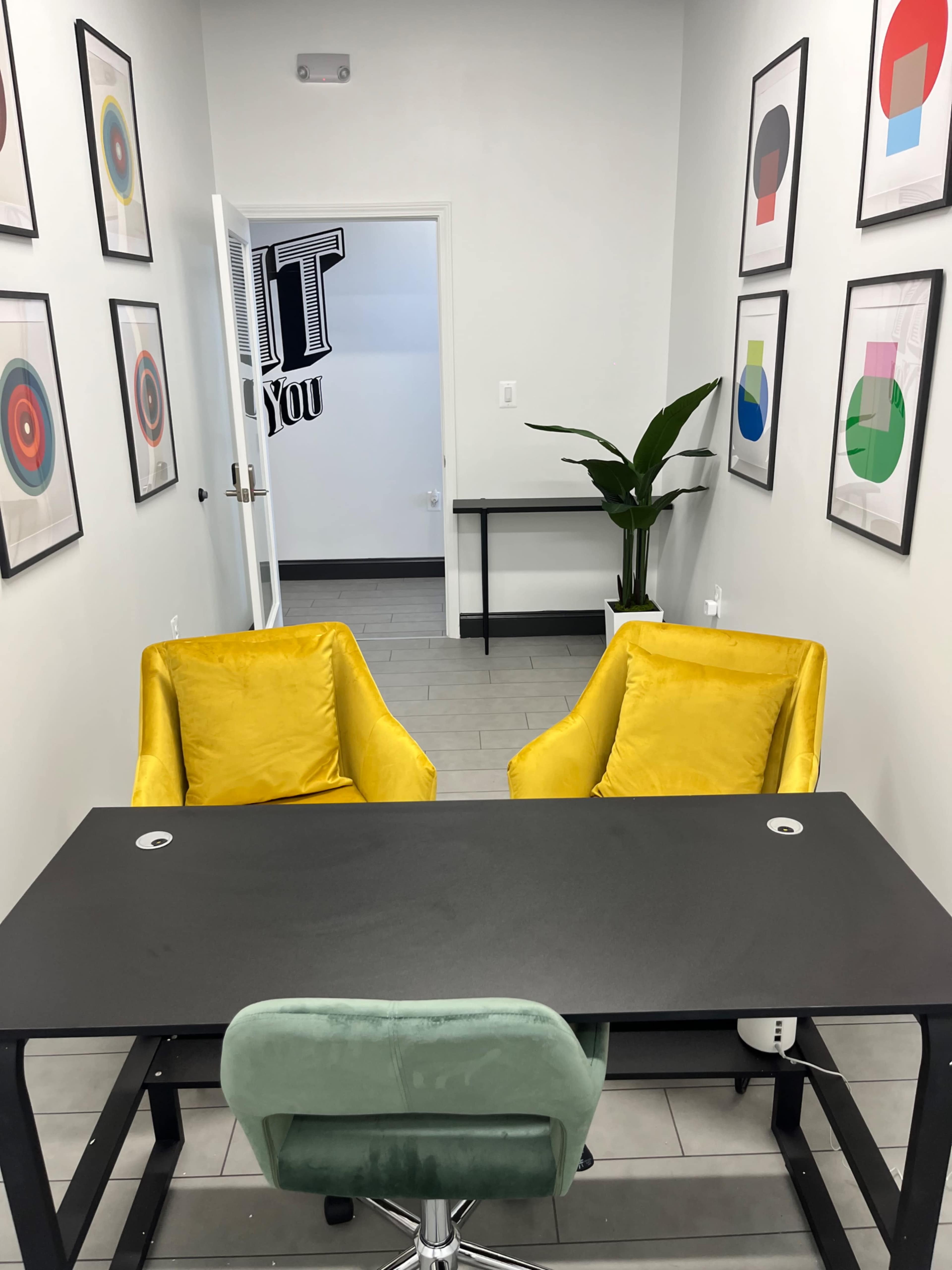 A small office features a black desk with two yellow armchairs and a green swivel chair, surrounded by framed abstract art on the walls.