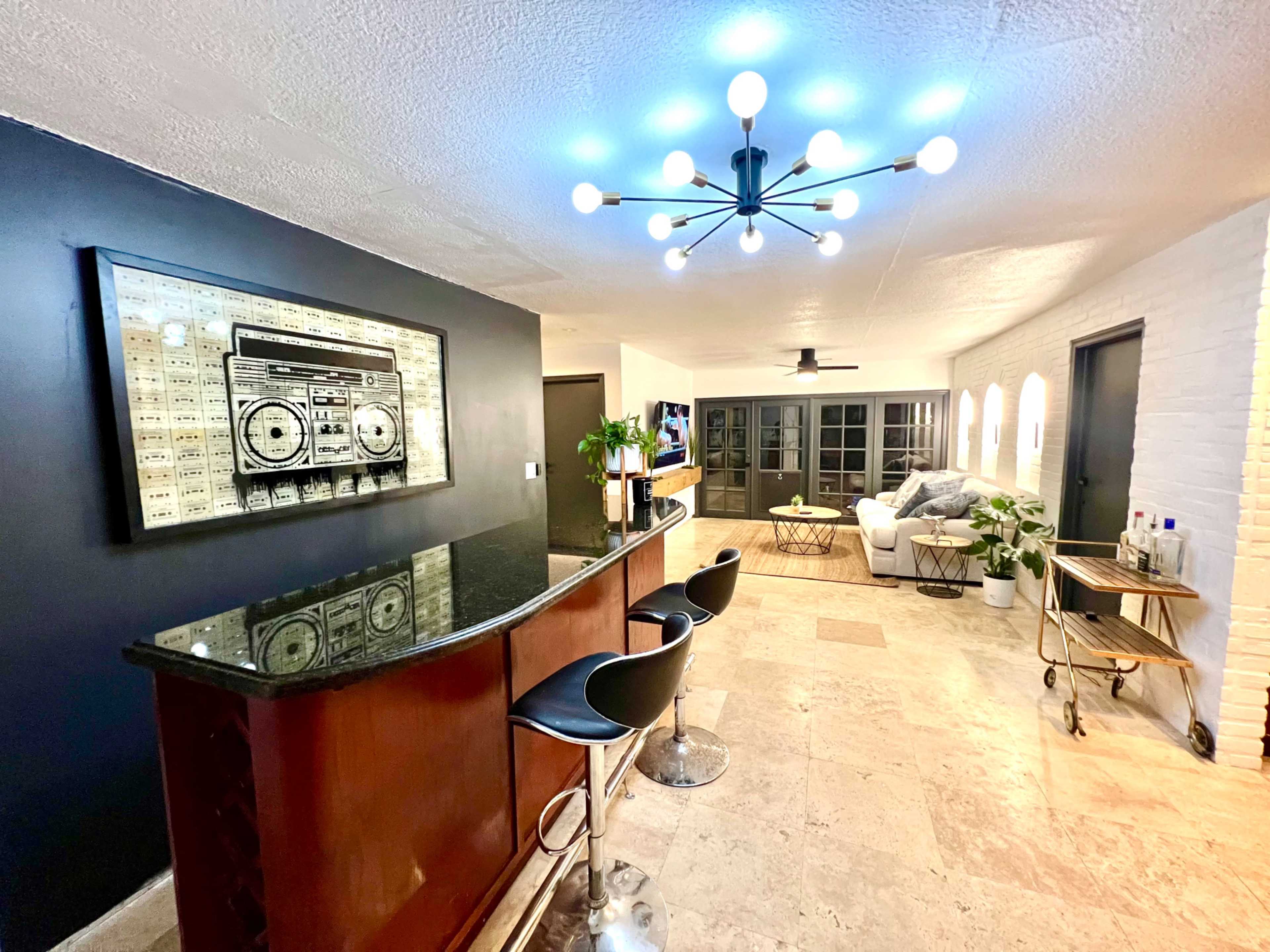 A modern living space features a bar with black stools, a stylish chandelier, and a framed artwork of a vintage boombox against a dark wall.