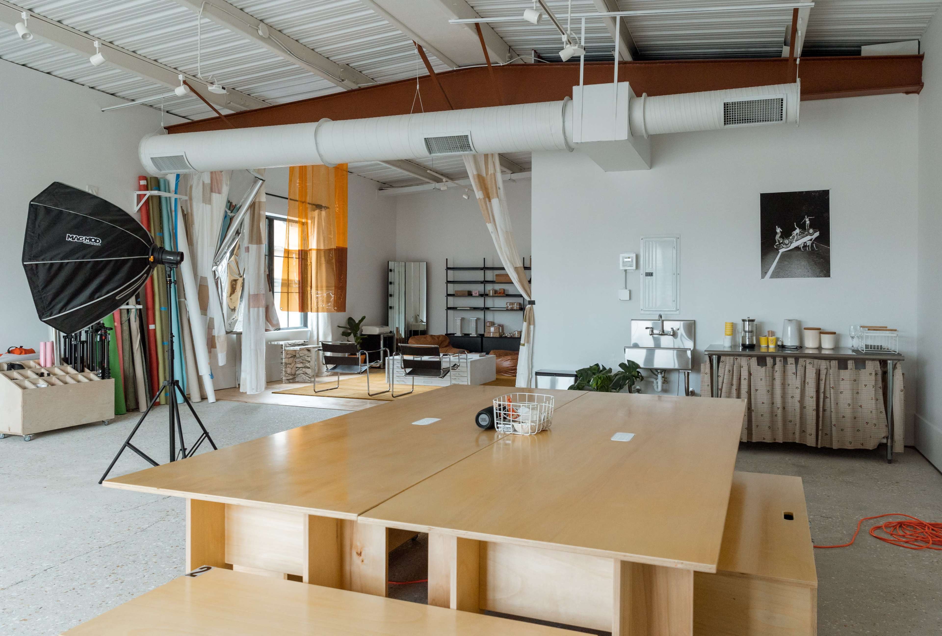 The image shows a spacious studio with a large wooden table, various lighting equipment, and a minimalist interior featuring shelves and couches.