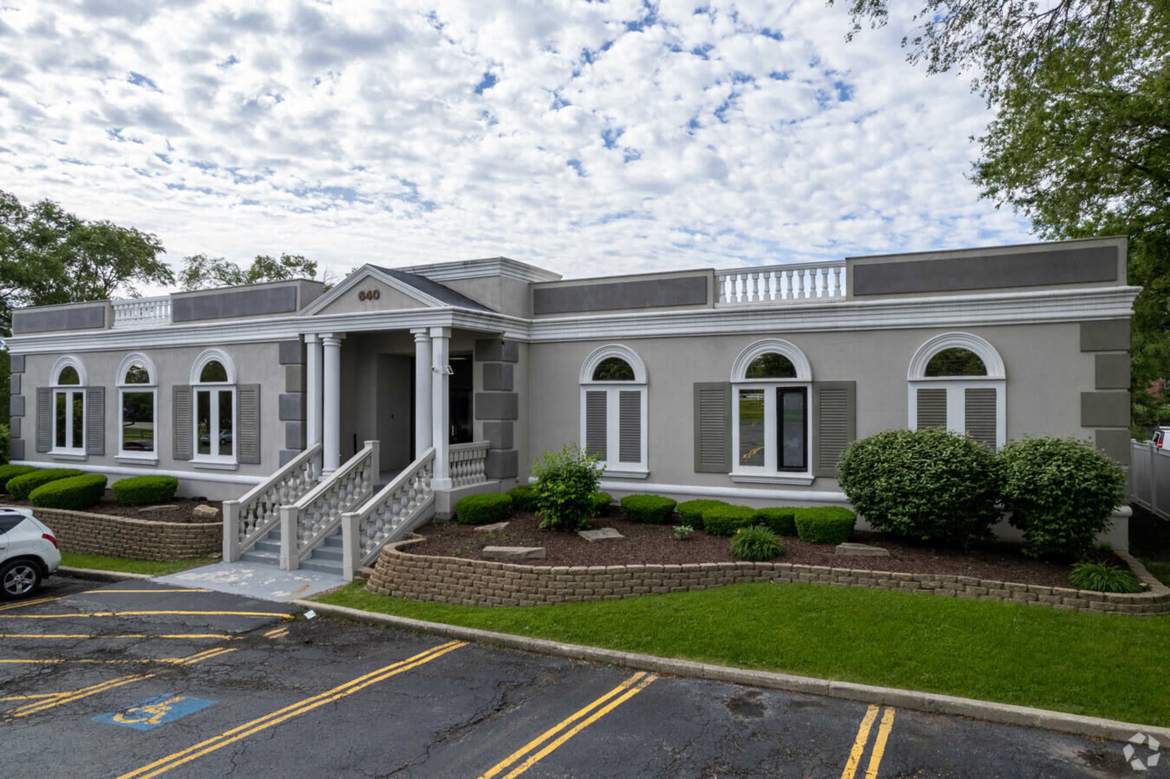 The image shows a single-story building with a symmetrical facade, featuring classical architectural details, a set of stairs leading to the entrance, and well-maintained landscaping.