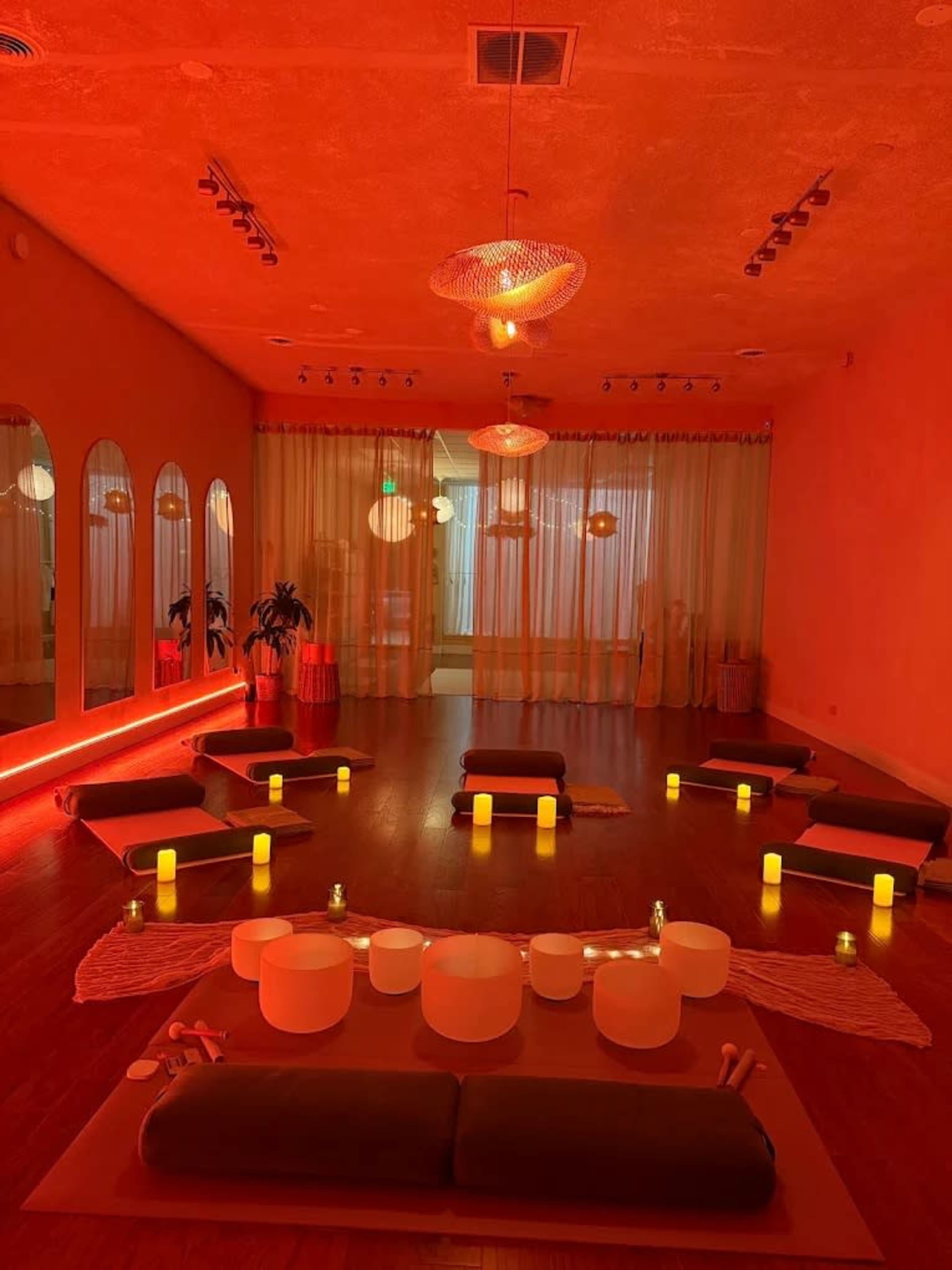 The image shows a tranquil meditation space with low seating cushions arranged in a circular pattern, illuminated by soft amber lighting and surrounded by candles.