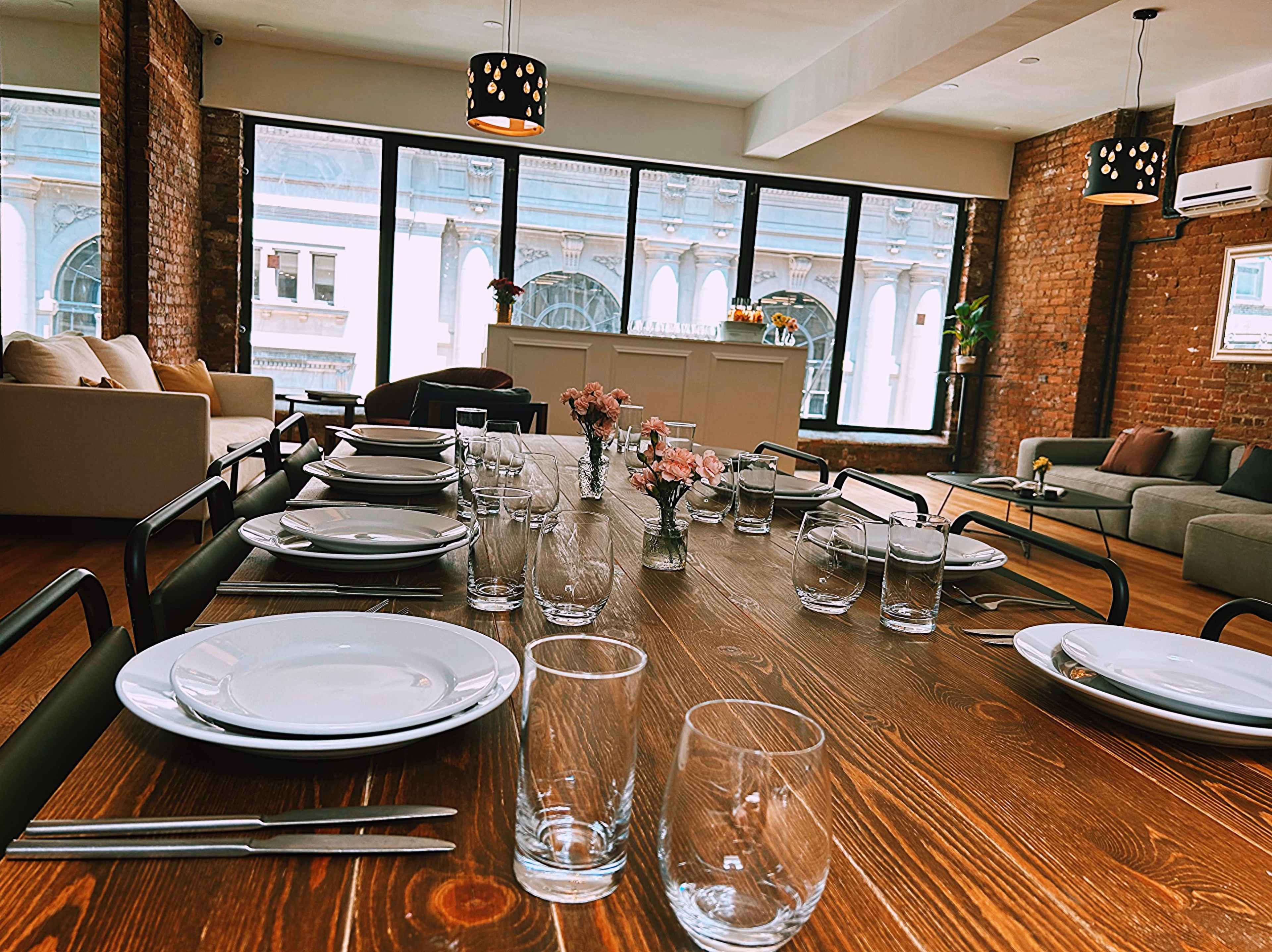 A long wooden dining table is set with plates, glasses, and small flower arrangements in a modern, well-lit room with large windows.