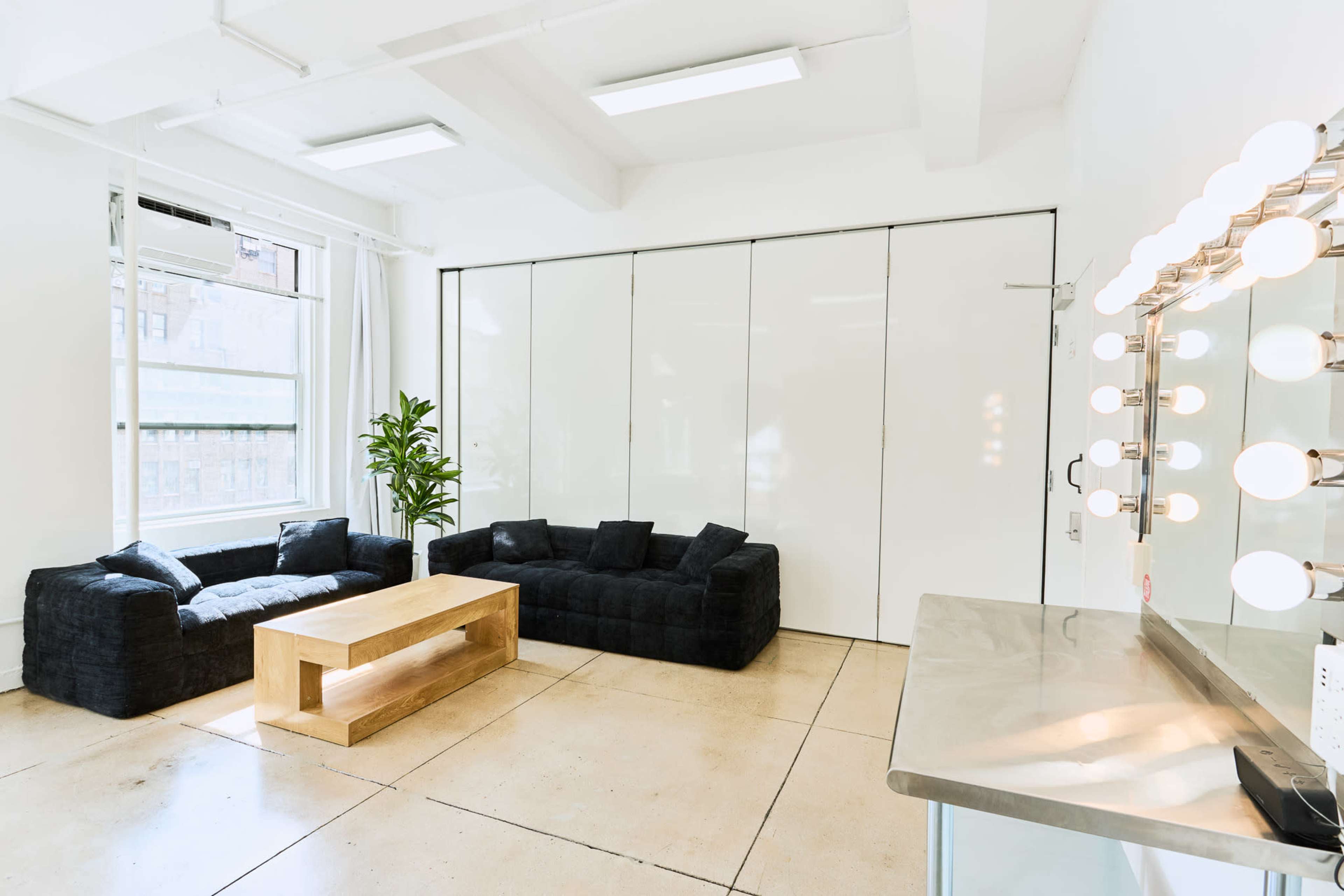 The image shows a minimalist interior space with two black sofas, a wooden coffee table, a plant, and a well-lit vanity mirror along one wall.