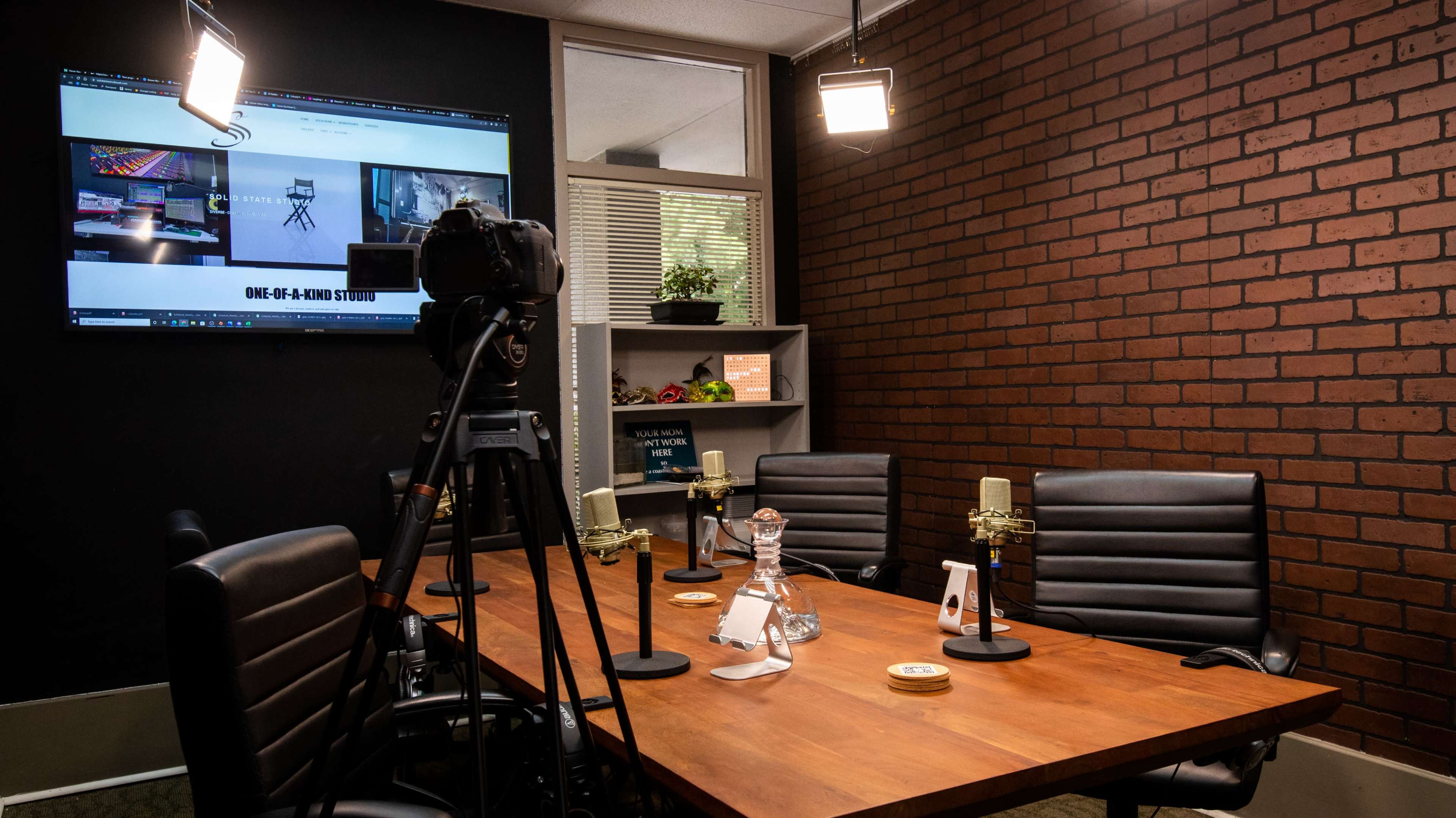 The image shows a video recording studio setup with a camera on a tripod, microphones on a wooden table, and a large screen displaying content on the wall.