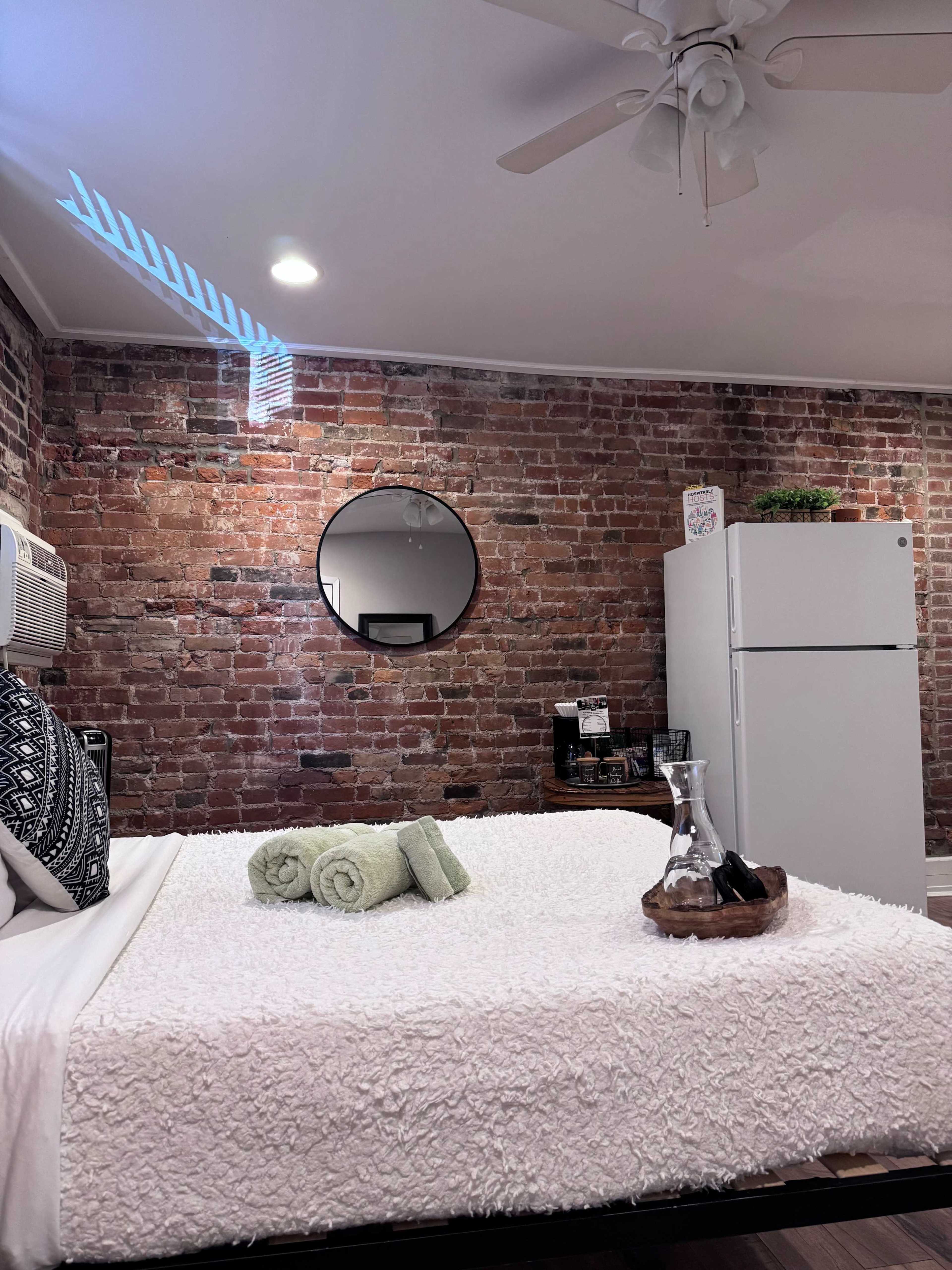 The image shows a cozy bedroom with exposed brick walls, a round mirror, a bed with white bedding, and a small refrigerator in the corner.