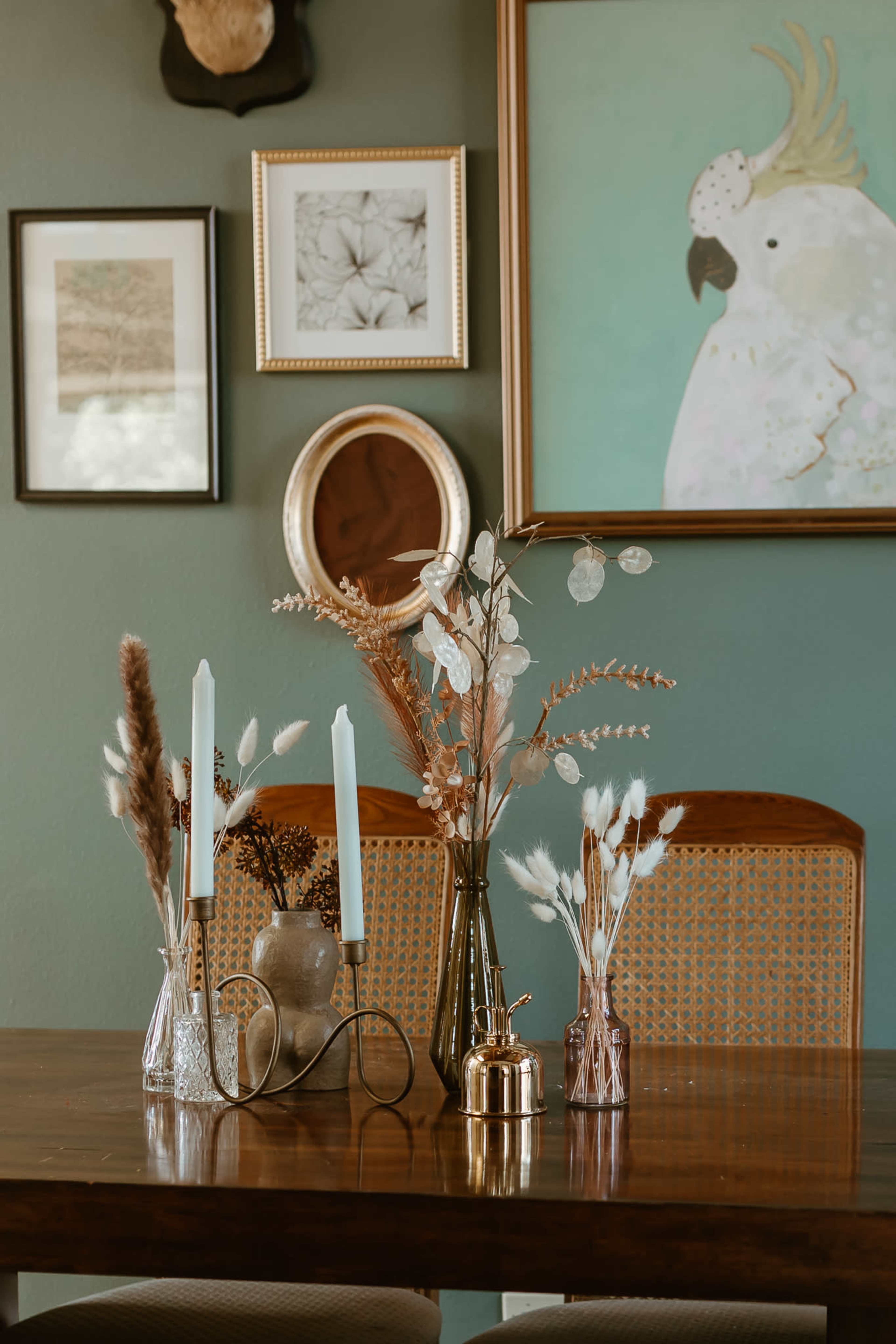 A dining table is decorated with various vases holding dried flowers and candles, surrounded by framed art on the green wall.