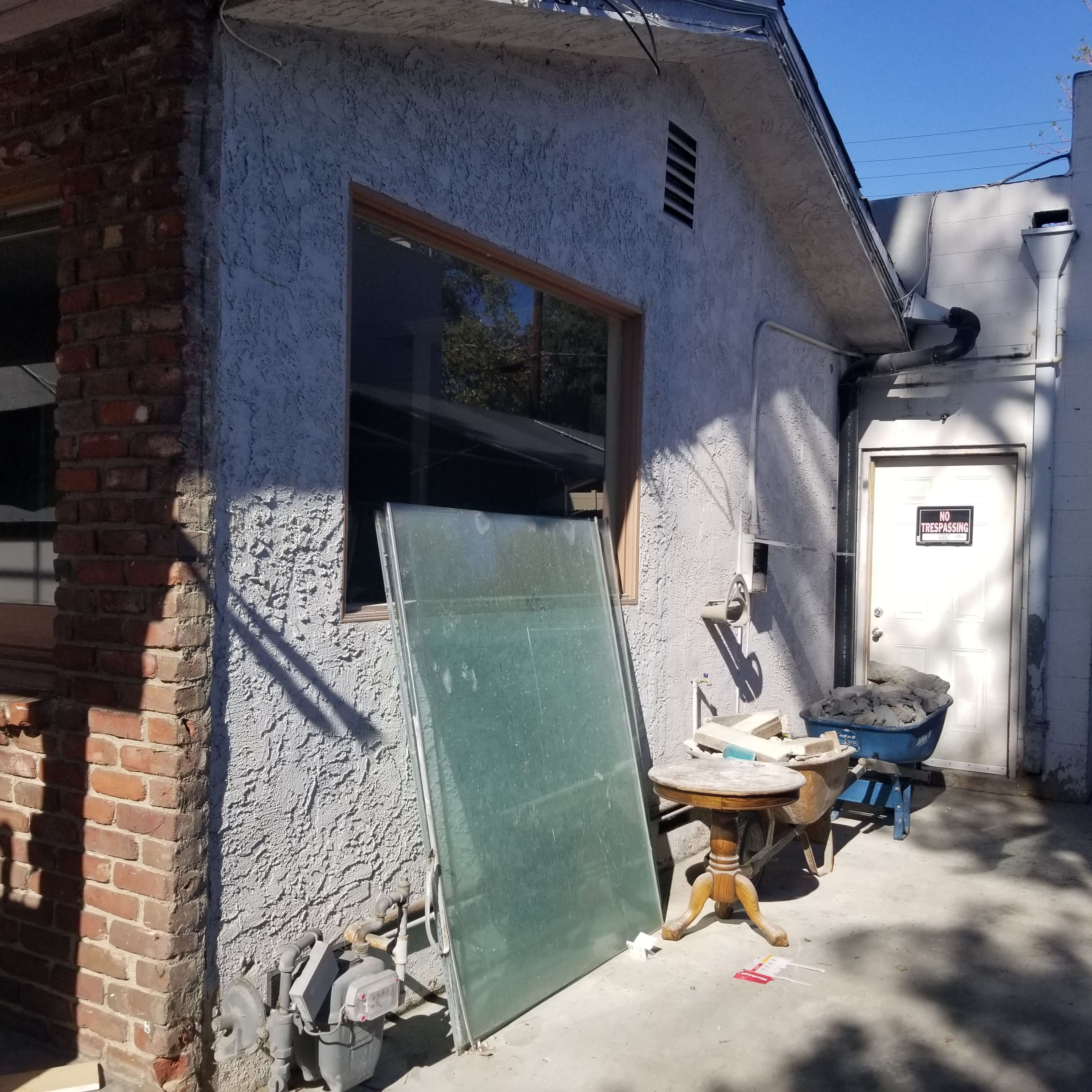 An exterior view of a building with a large glass panel leaning against the wall, a wooden table, and a door marked with a sign.