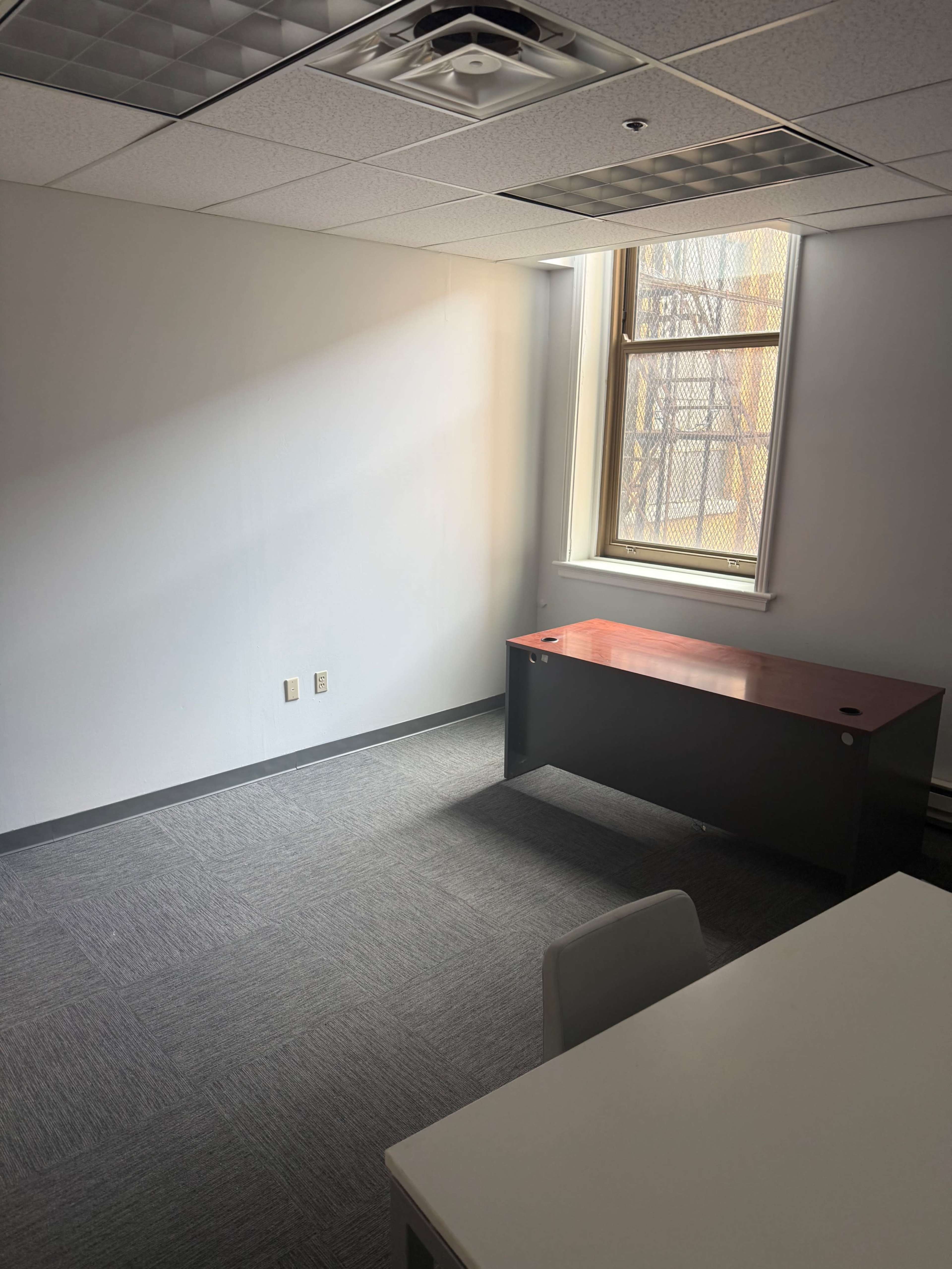 The image shows a sparsely furnished office with a wooden desk, a white table, and a window allowing natural light to enter.