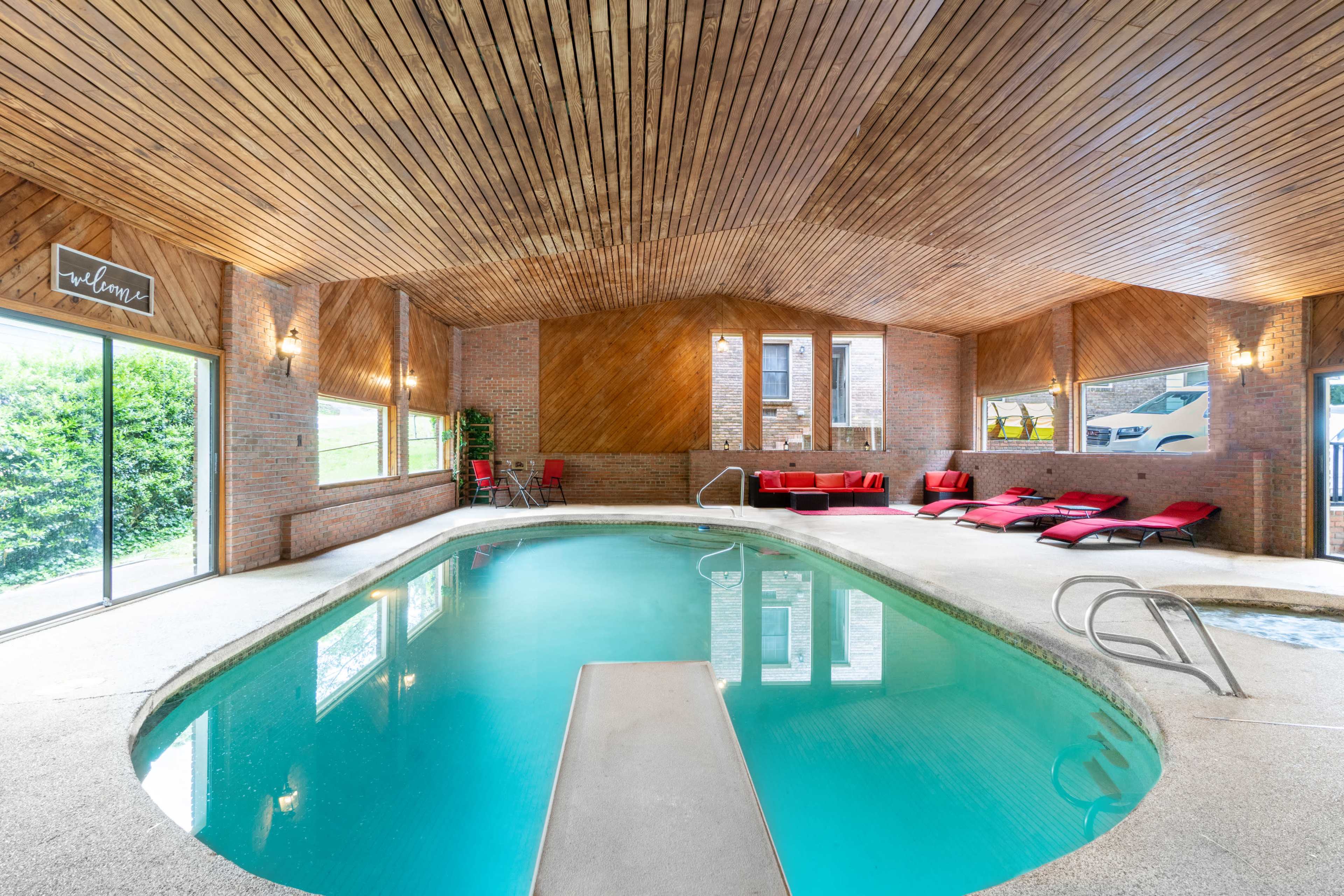 The image shows an indoor swimming pool area with a wooden ceiling, stone walls, and several red lounge chairs beside the pool.