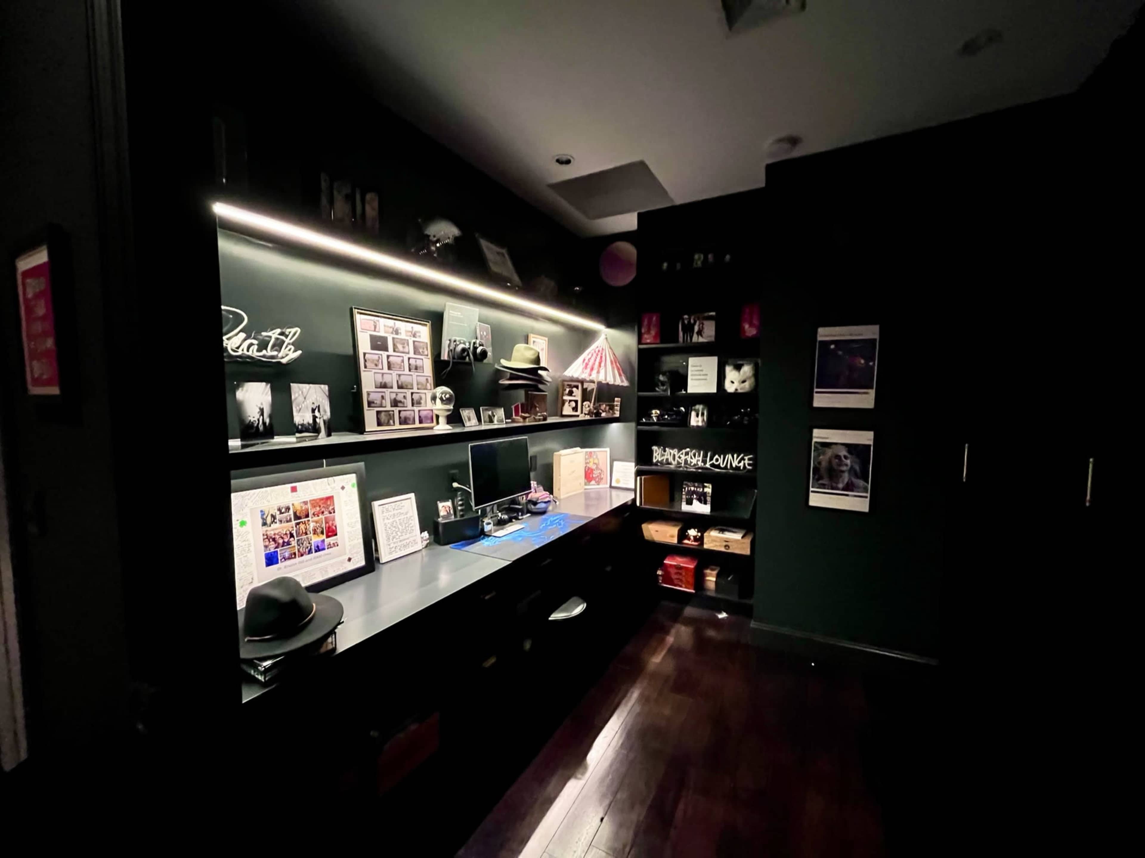 The image shows a dark home office featuring a long desk with a computer, shelves lined with photos and decorative items, and a wall with framed pictures.