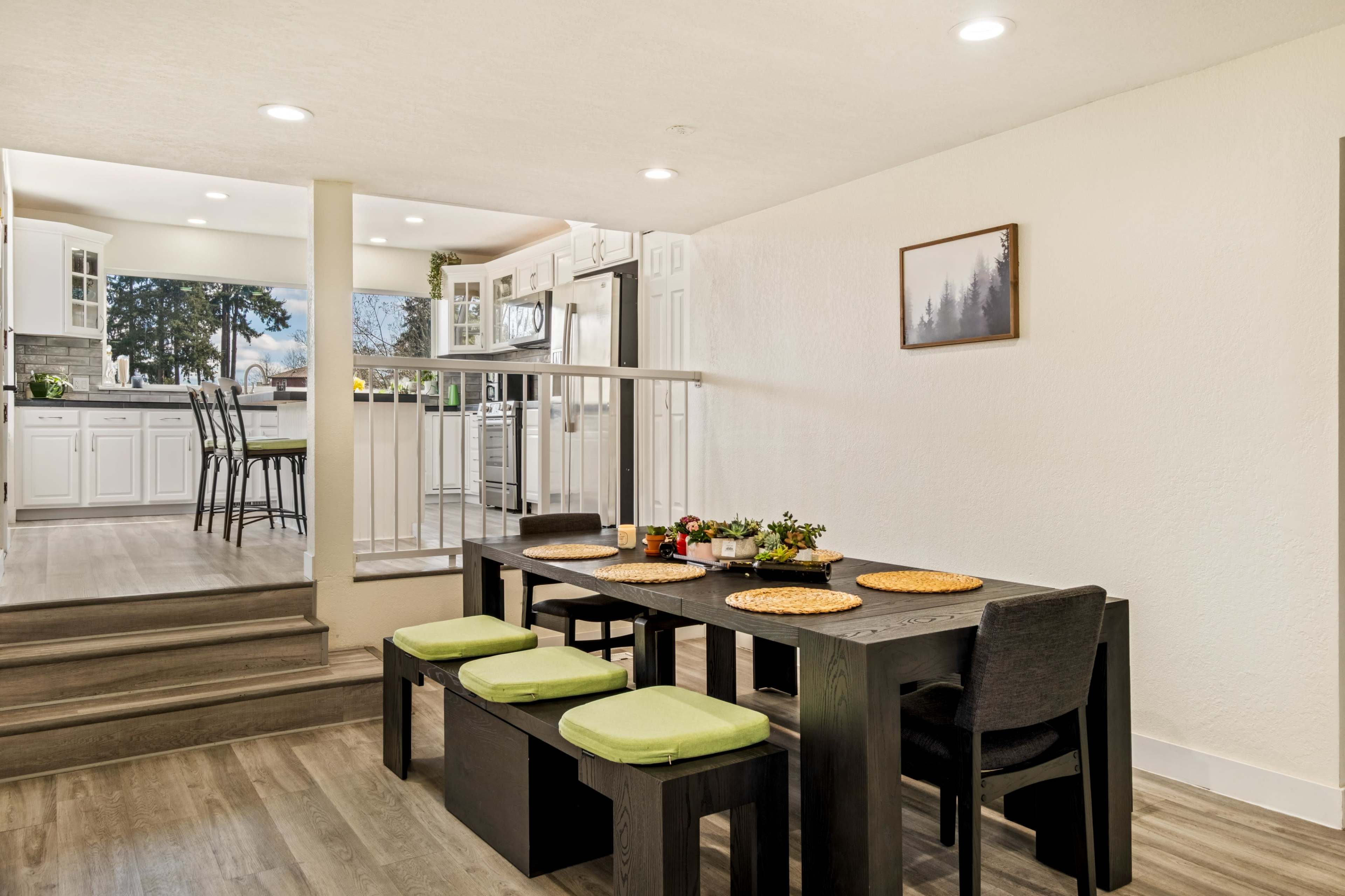 The image shows a dining area with a long black table and green cushions, adjacent to a modern kitchen visible in the background.