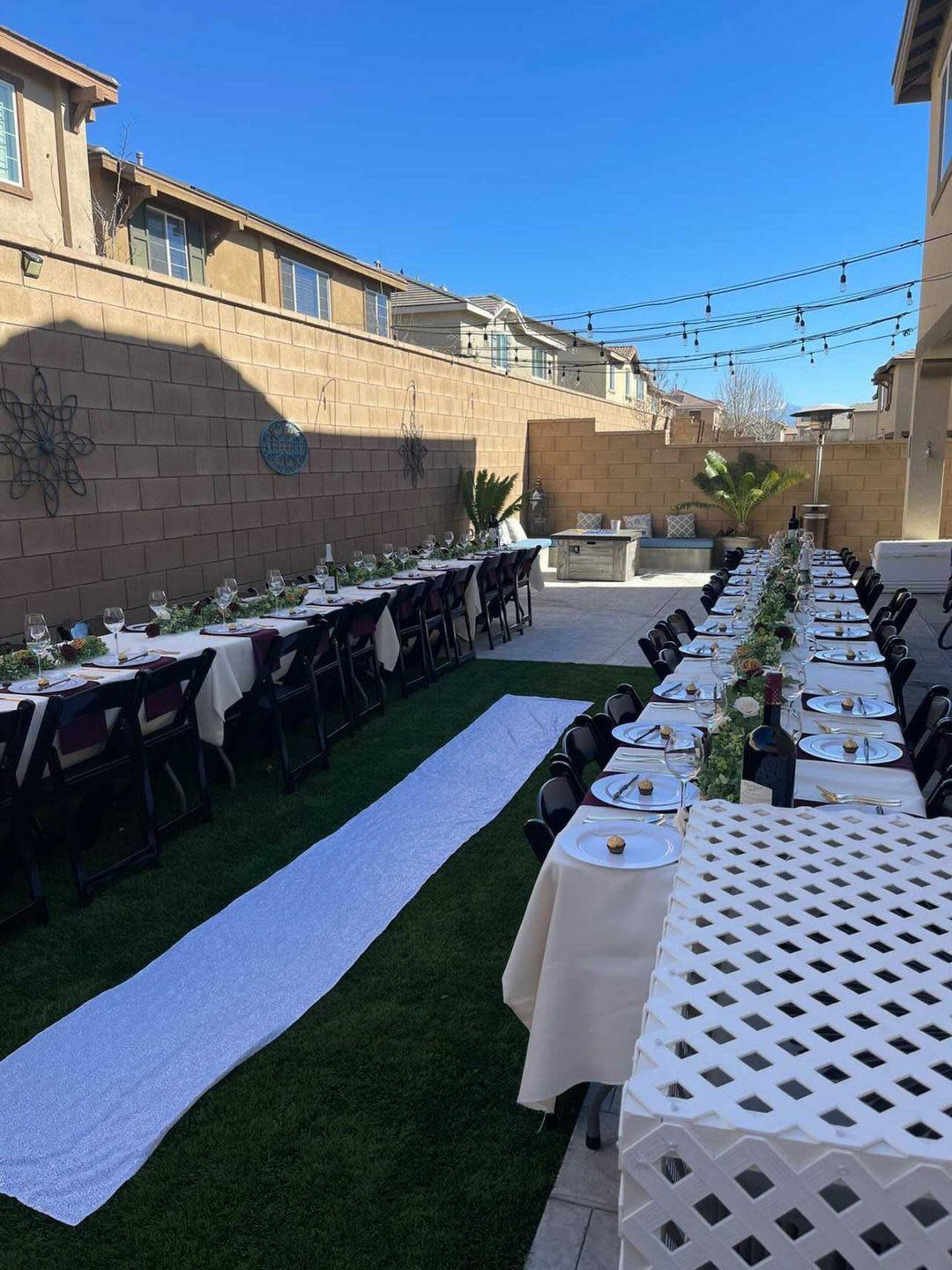 A long outdoor dining setup with neatly arranged tables on grass, flanked by a white pathway and surrounded by a decorative wall and houses.