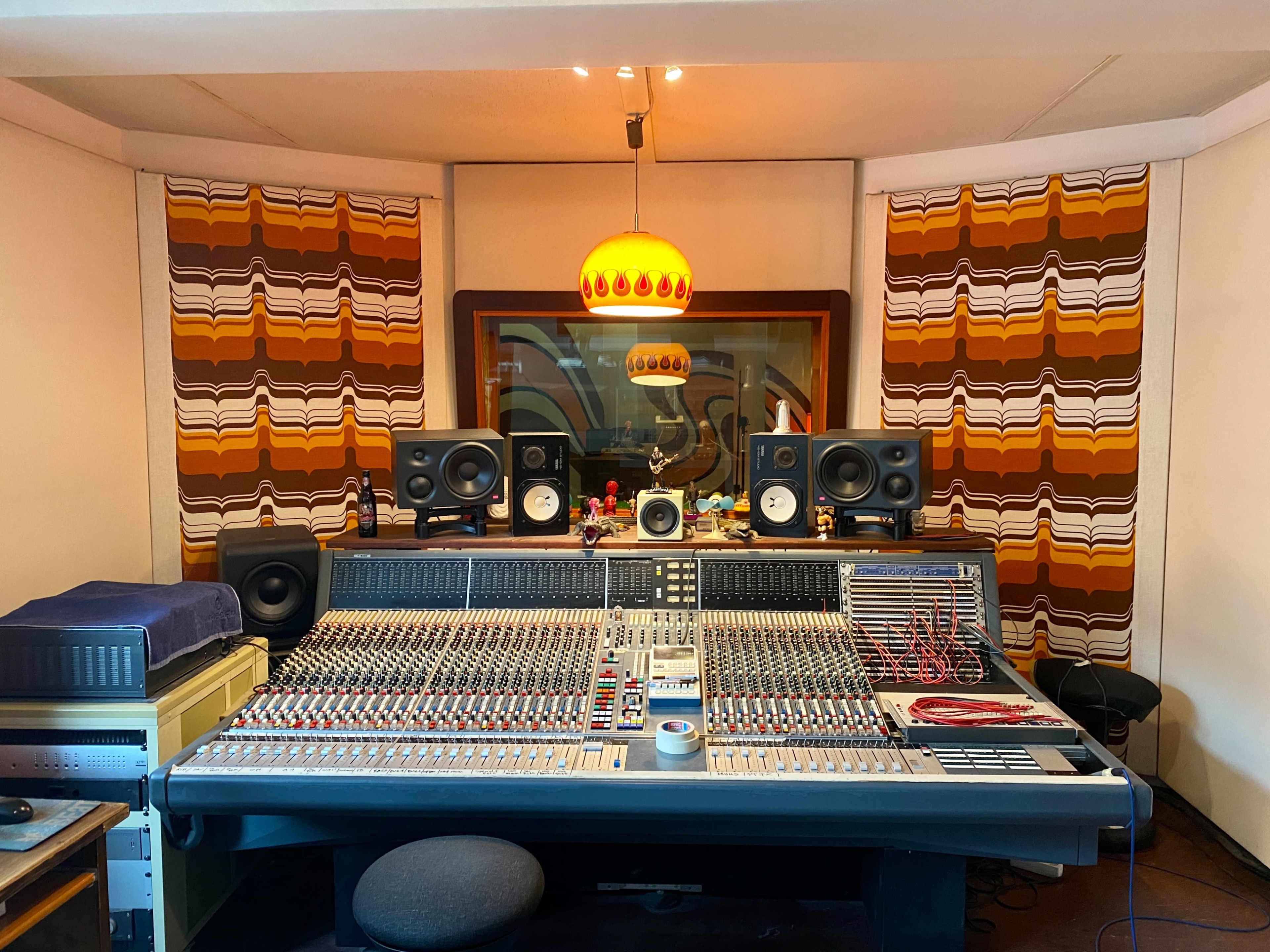 A recording studio with a large mixing console at the center, flanked by tall speakers and decorated with retro-patterned curtains.