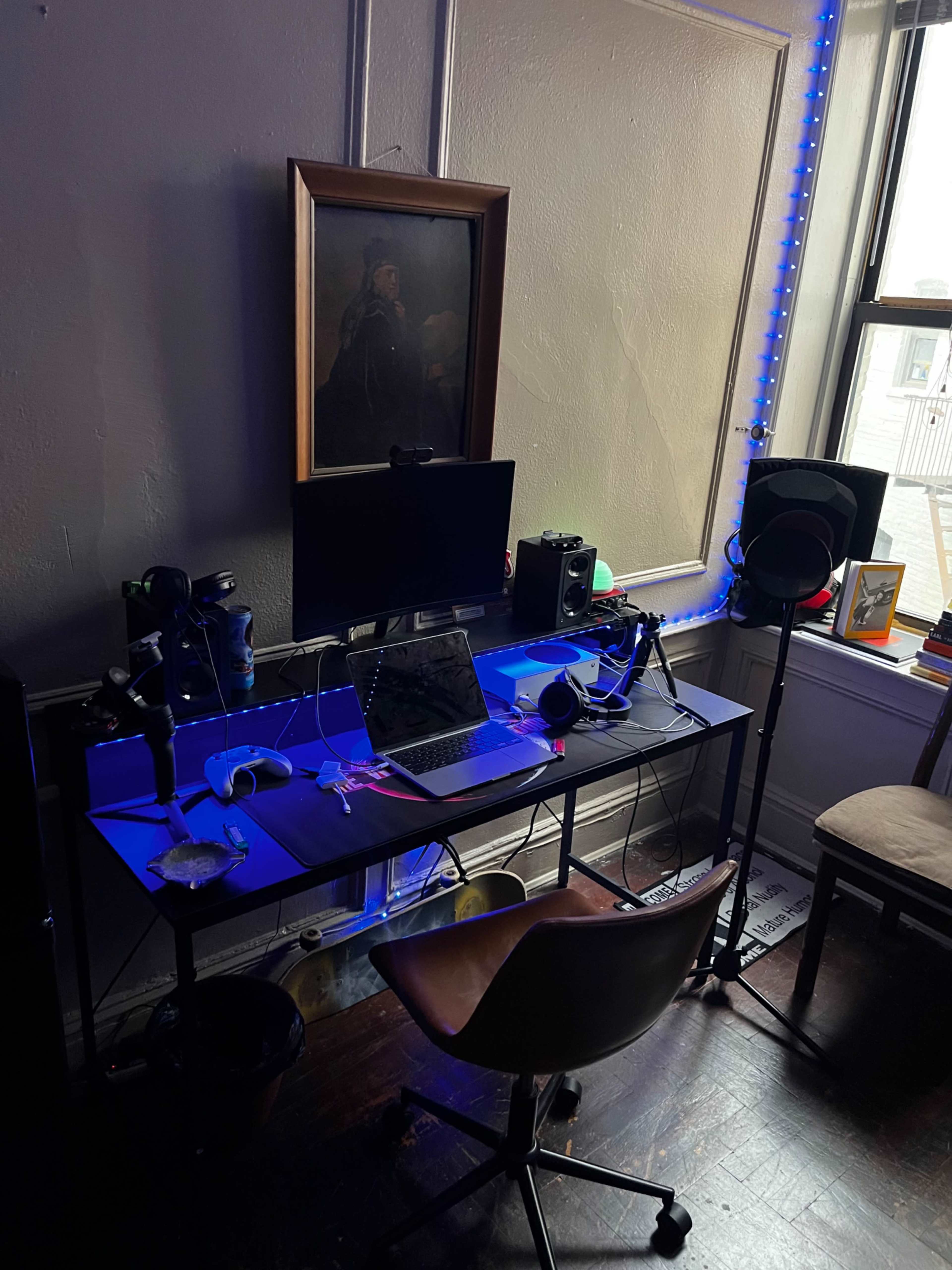A desk setup featuring a computer monitor, a laptop, audio equipment, and a chair, positioned near a window with blue LED lights and a framed artwork on the wall.
