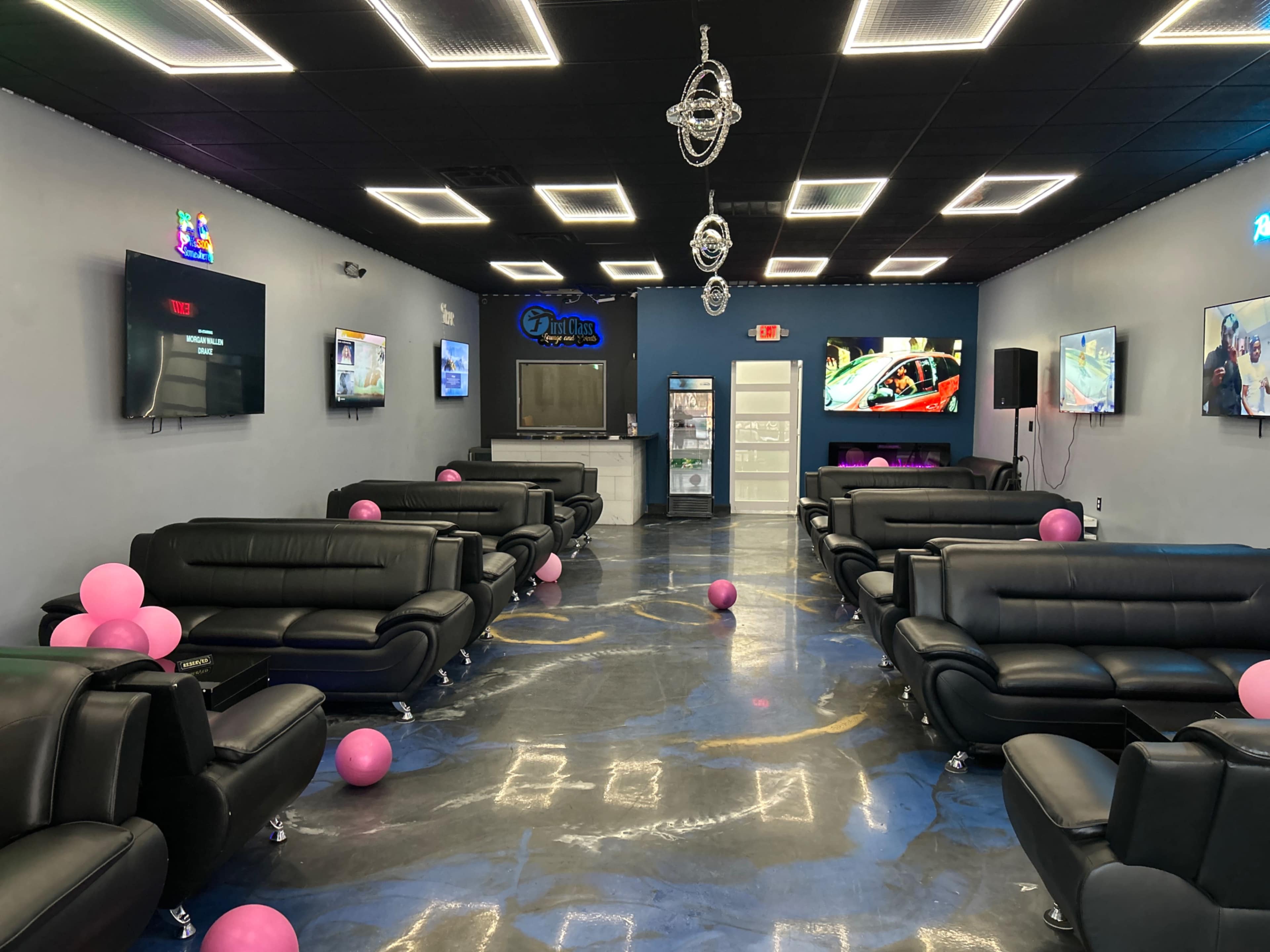 A modern lounge area with black leather sofas, several flat-screen TVs on the walls, and pink balloons scattered across the floor.