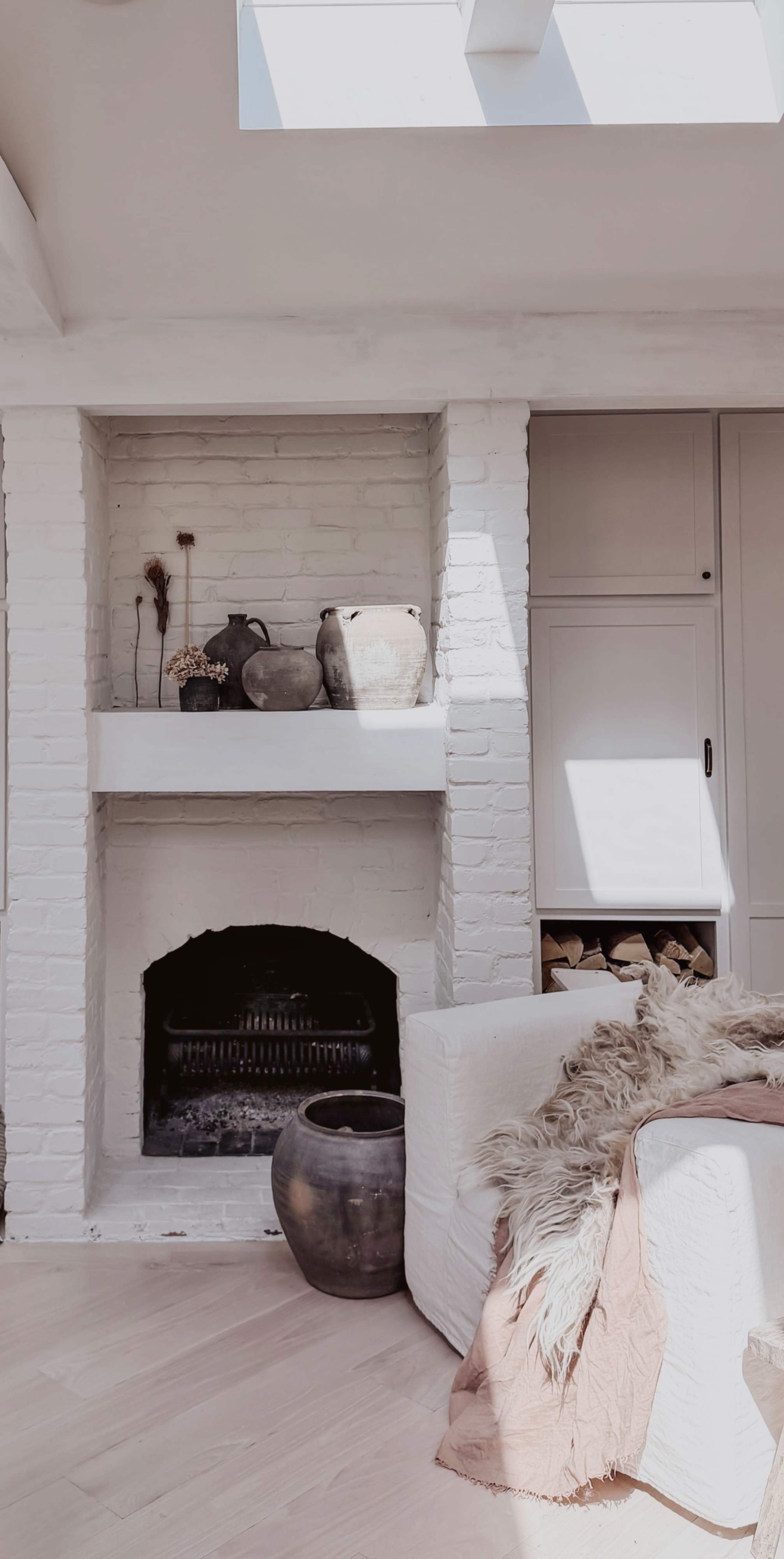The image shows a living room interior featuring a white brick fireplace with decorative vases on a shelf, a cozy sofa with a throw blanket, and storage cabinets beside the fireplace.