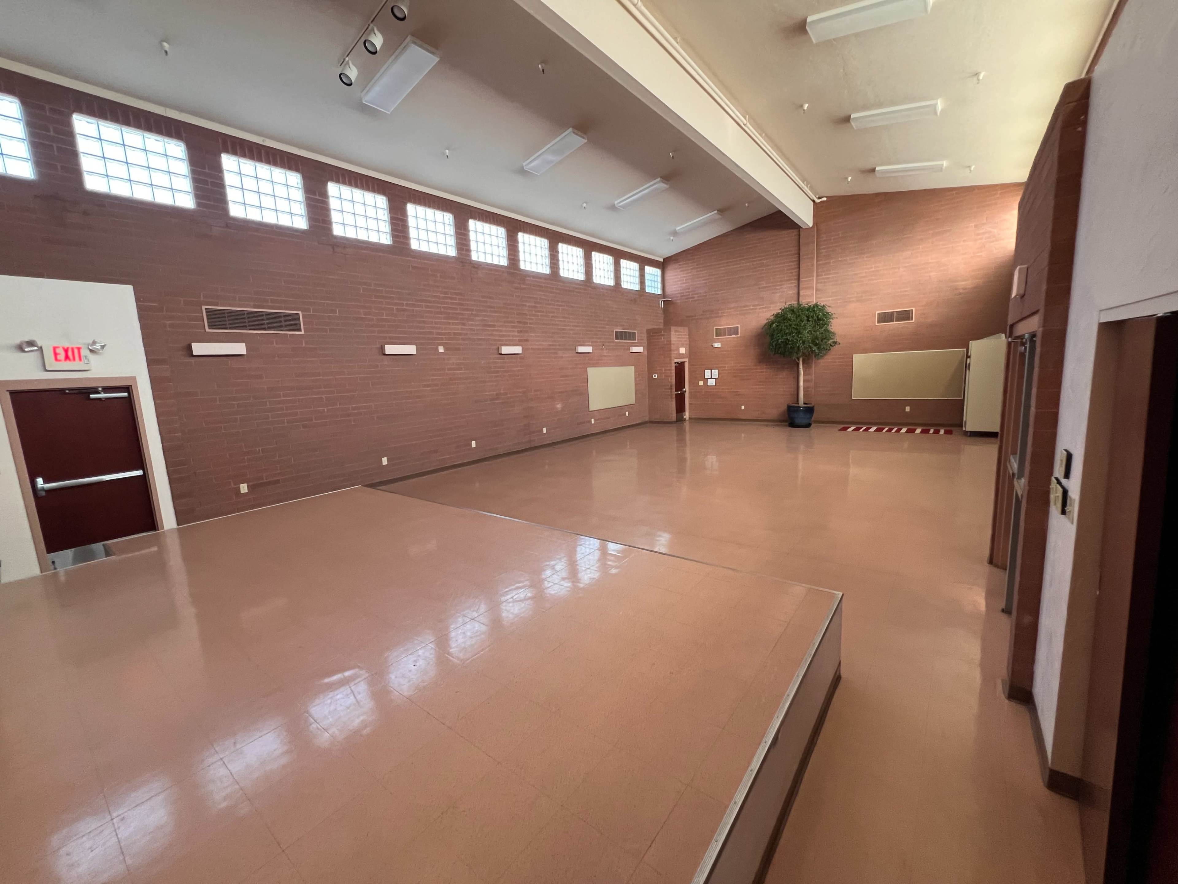 The image shows a spacious, empty room with brick walls, large windows, and wooden flooring.