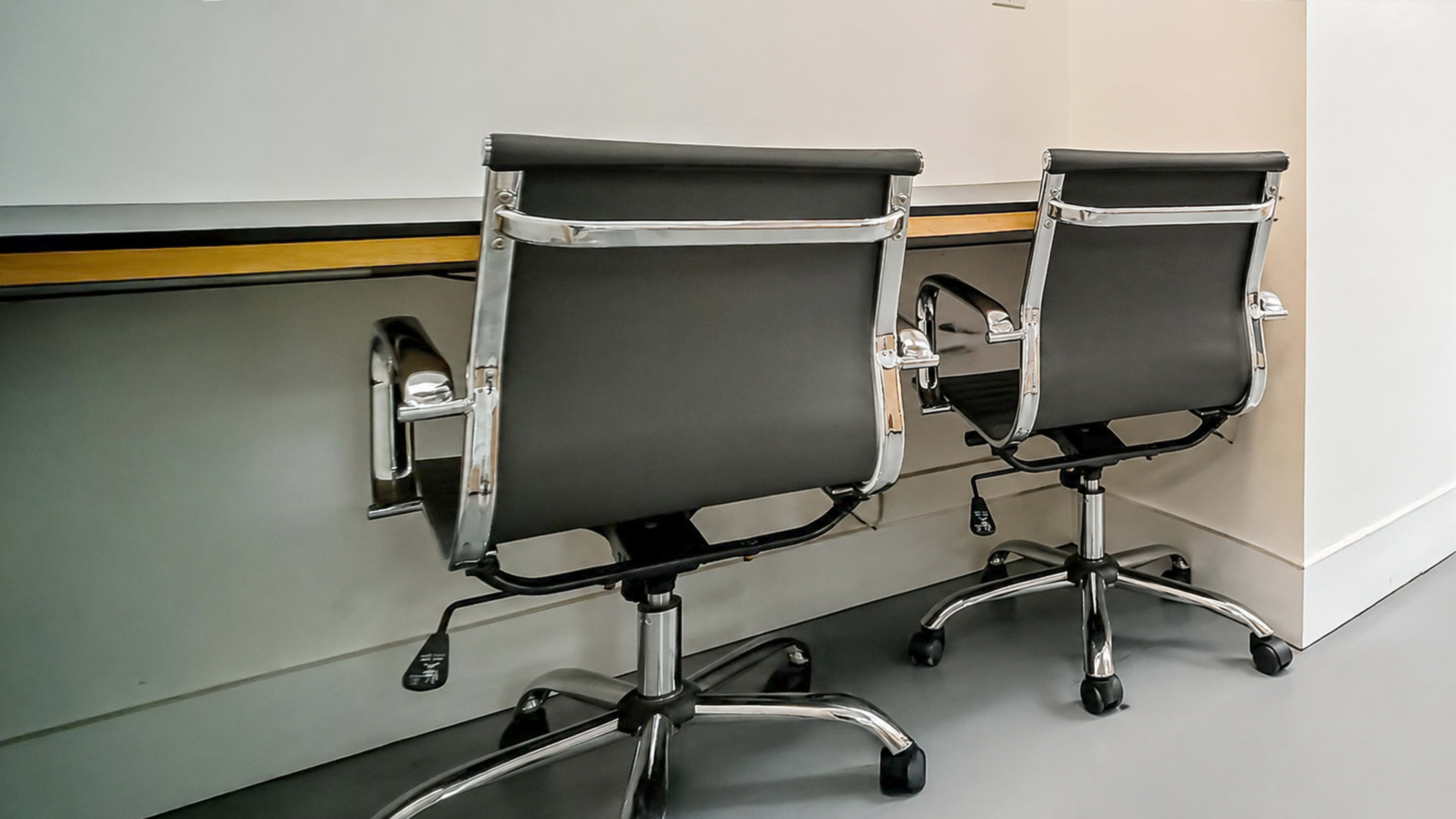 Two office chairs with chrome frames and black upholstery are positioned in front of a simple desk against a wall.