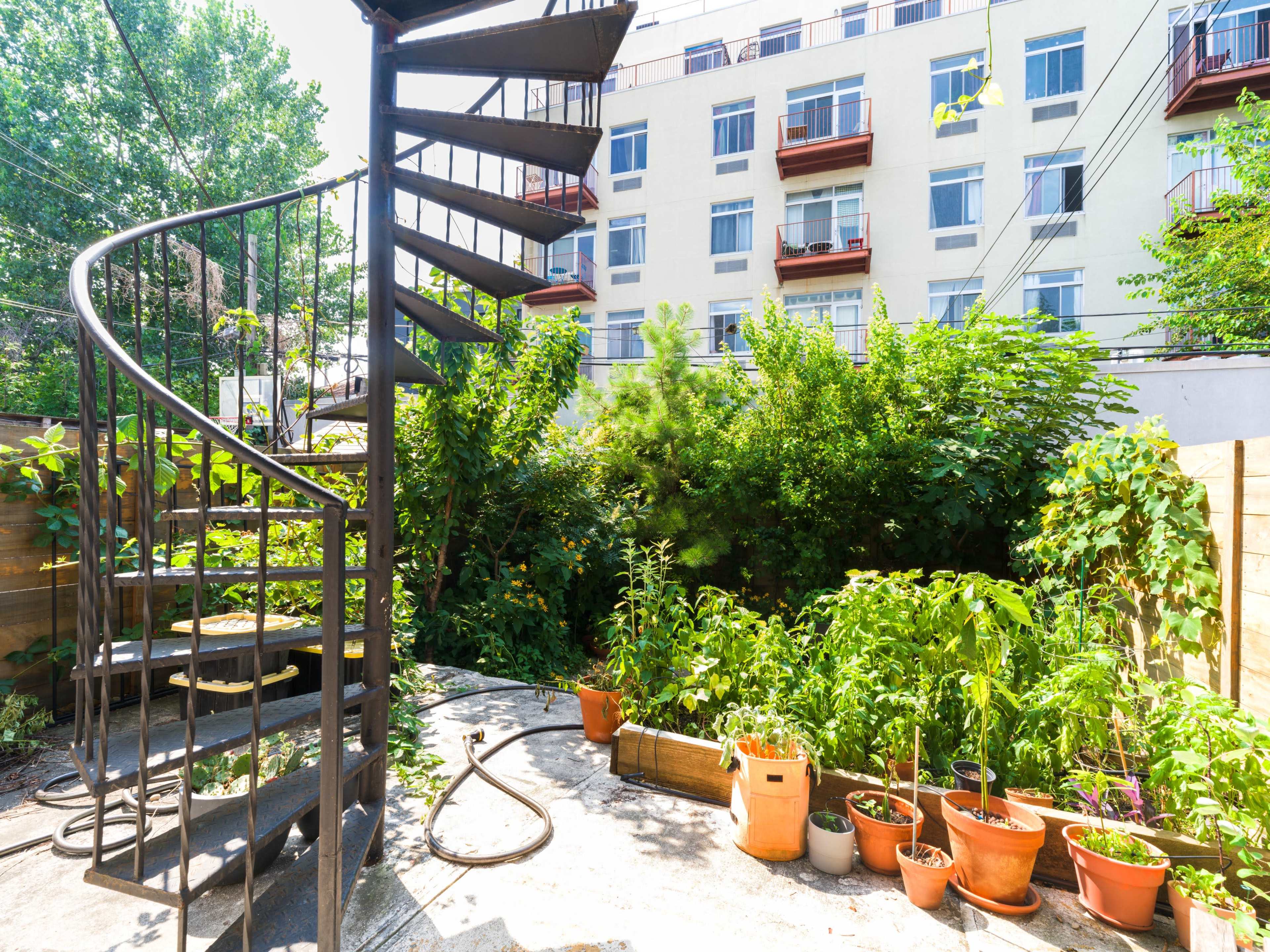 A spiral staircase leads from a concrete patio surrounded by lush greenery and potted plants to an upper floor of a building.