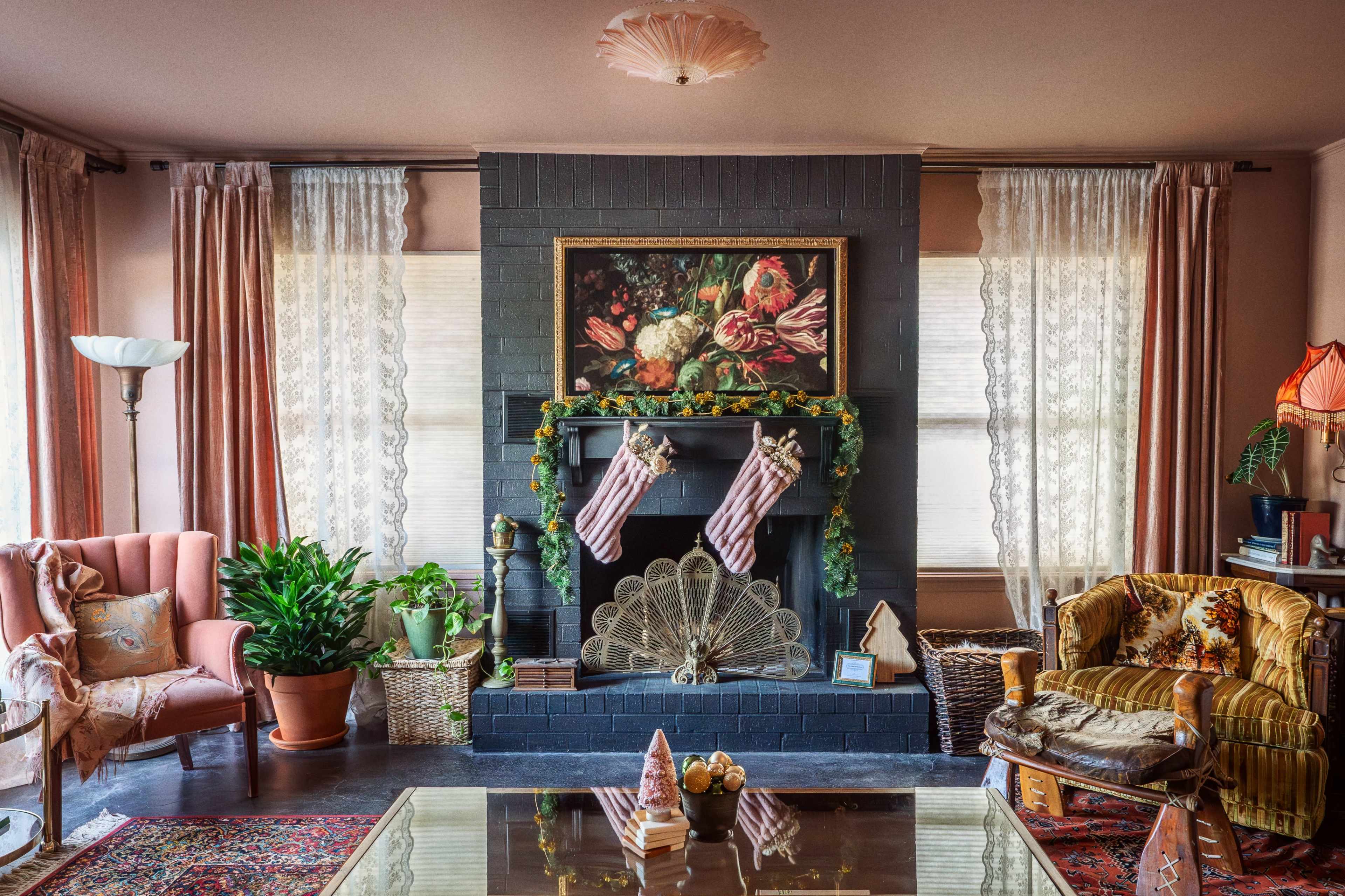 The living room features a fireplace adorned with stockings, a floral painting, and decorated with plants and furniture in warm colors.