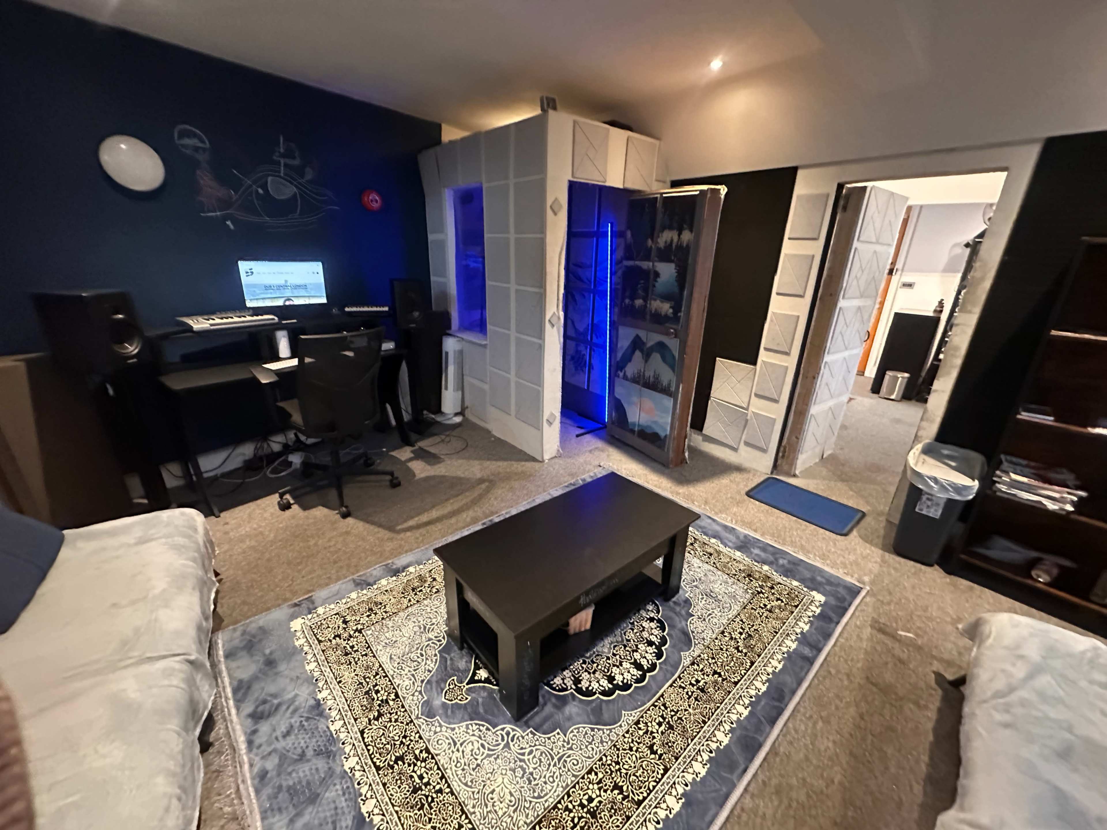 The image shows a room featuring a desk with a computer setup, a cozy seating area with sofas, and a patterned rug on the floor.